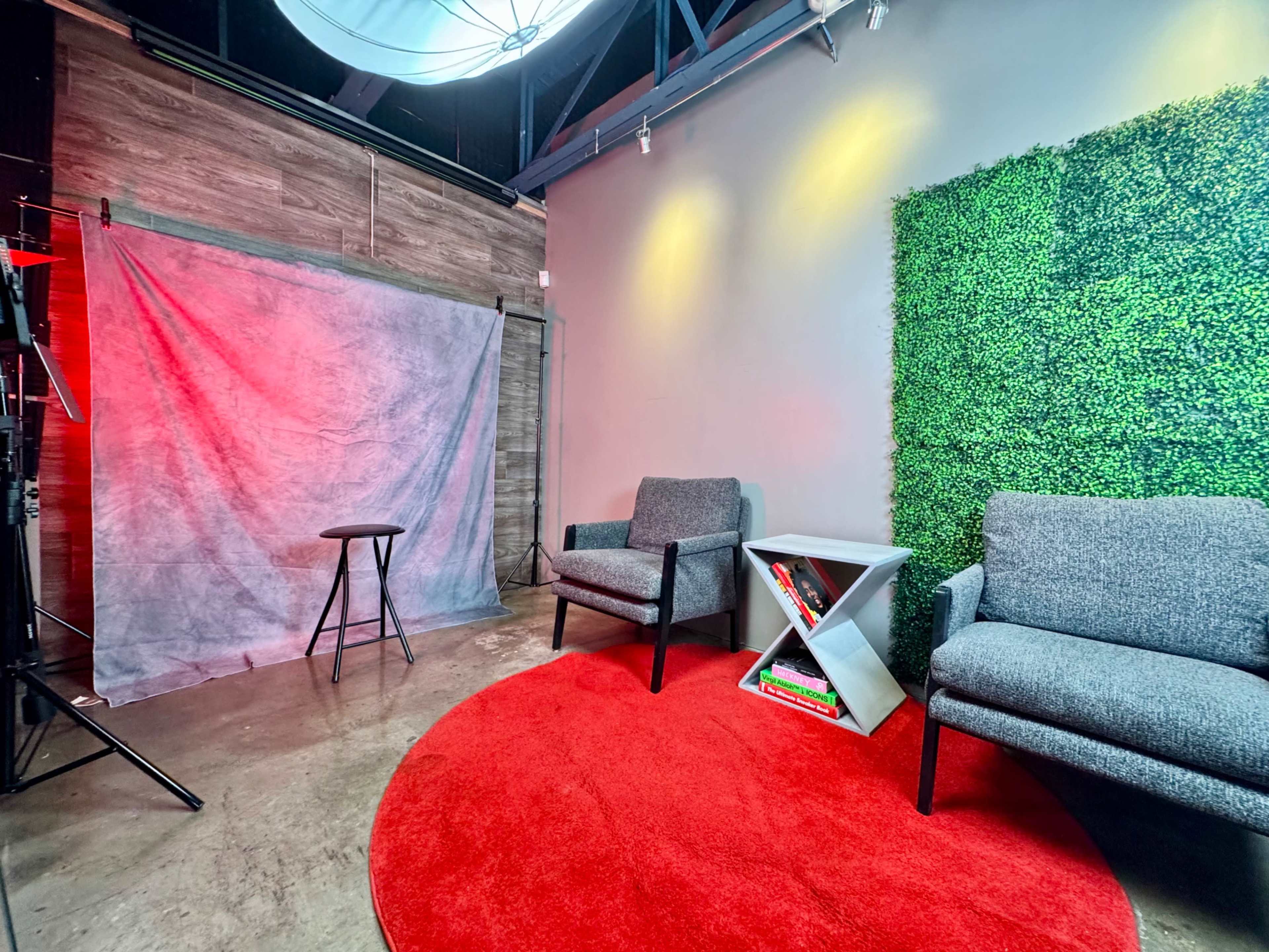 The image shows a small studio space featuring two gray chairs on a red circular rug, a white side table, a stool, and a backdrop with a green wall and gray fabric.