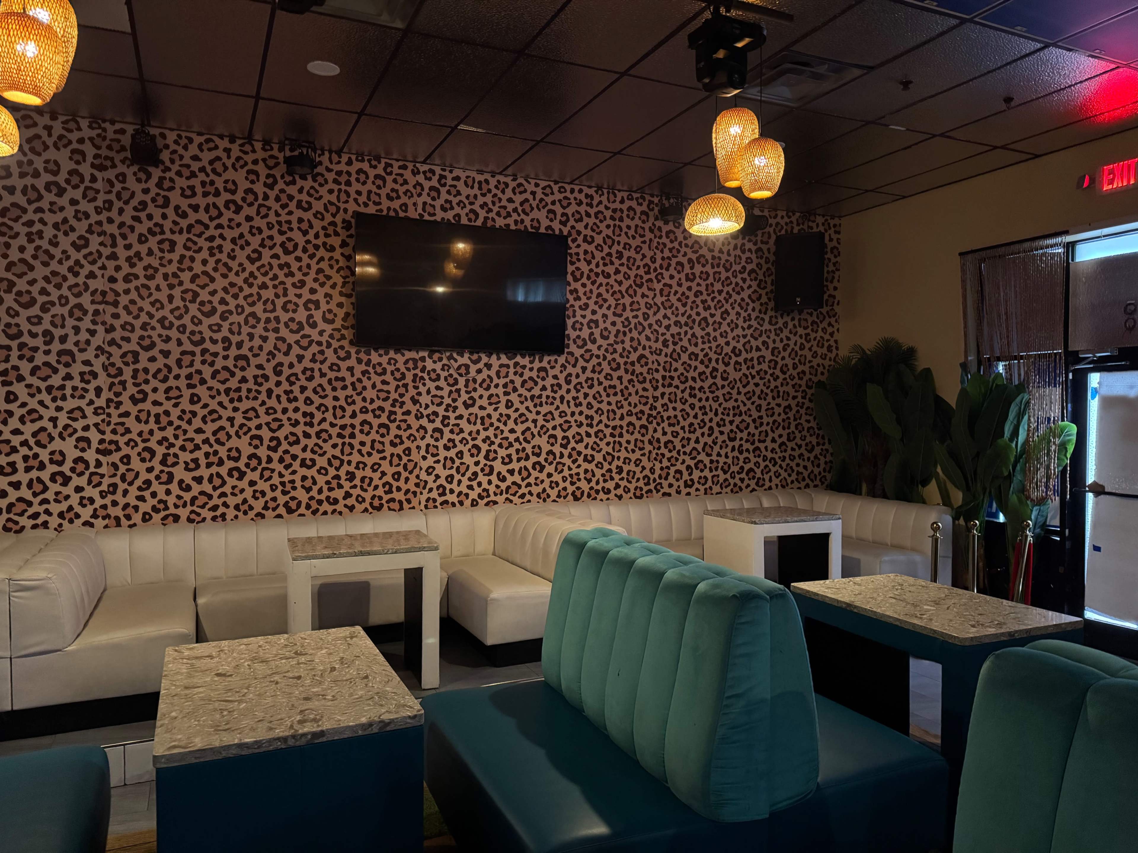 The image shows a modern lounge with white leather banquettes, teal seating, and a leopard print accent wall, featuring a large flat-screen television.