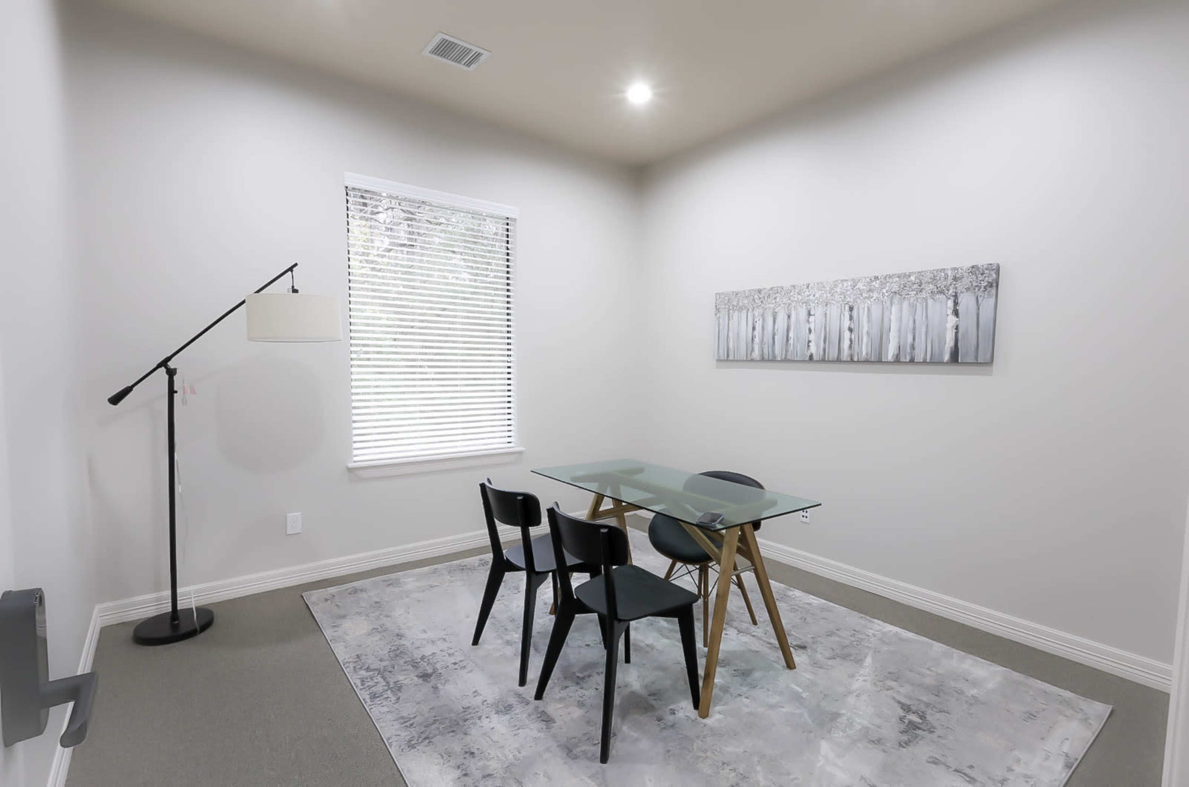 A minimalist room features a glass dining table with four black chairs, a floor lamp, and a wall art piece depicting birch trees, all set against a light-colored backdrop.