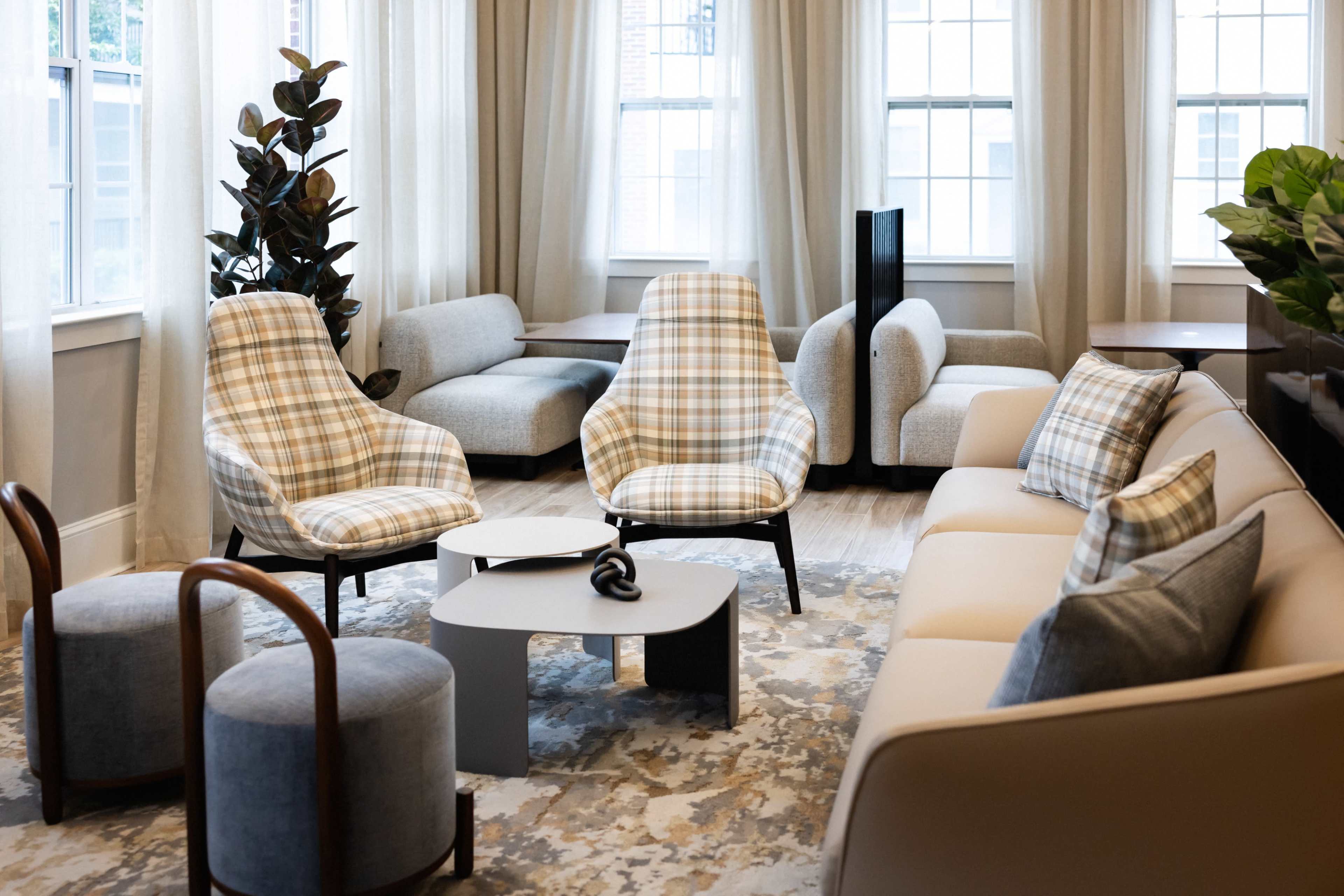 The image shows a modern lounge area featuring patterned armchairs, a light-colored sofa, a round coffee table, and potted plants, all arranged on a textured rug.