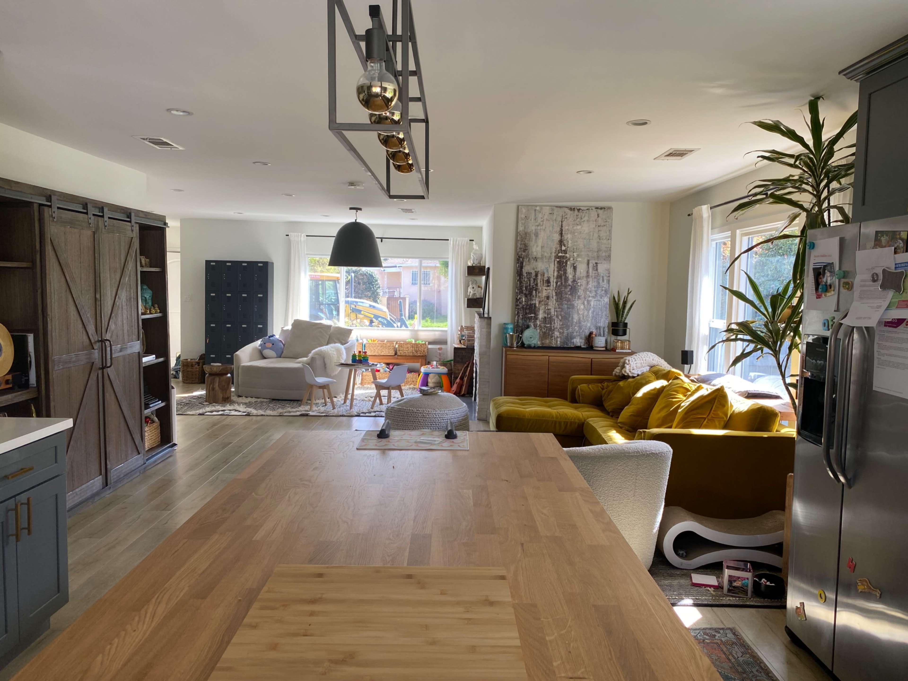 A modern kitchen and living area with an island, sunlight streaming through large windows, and a mix of furniture including a couch and armchairs.