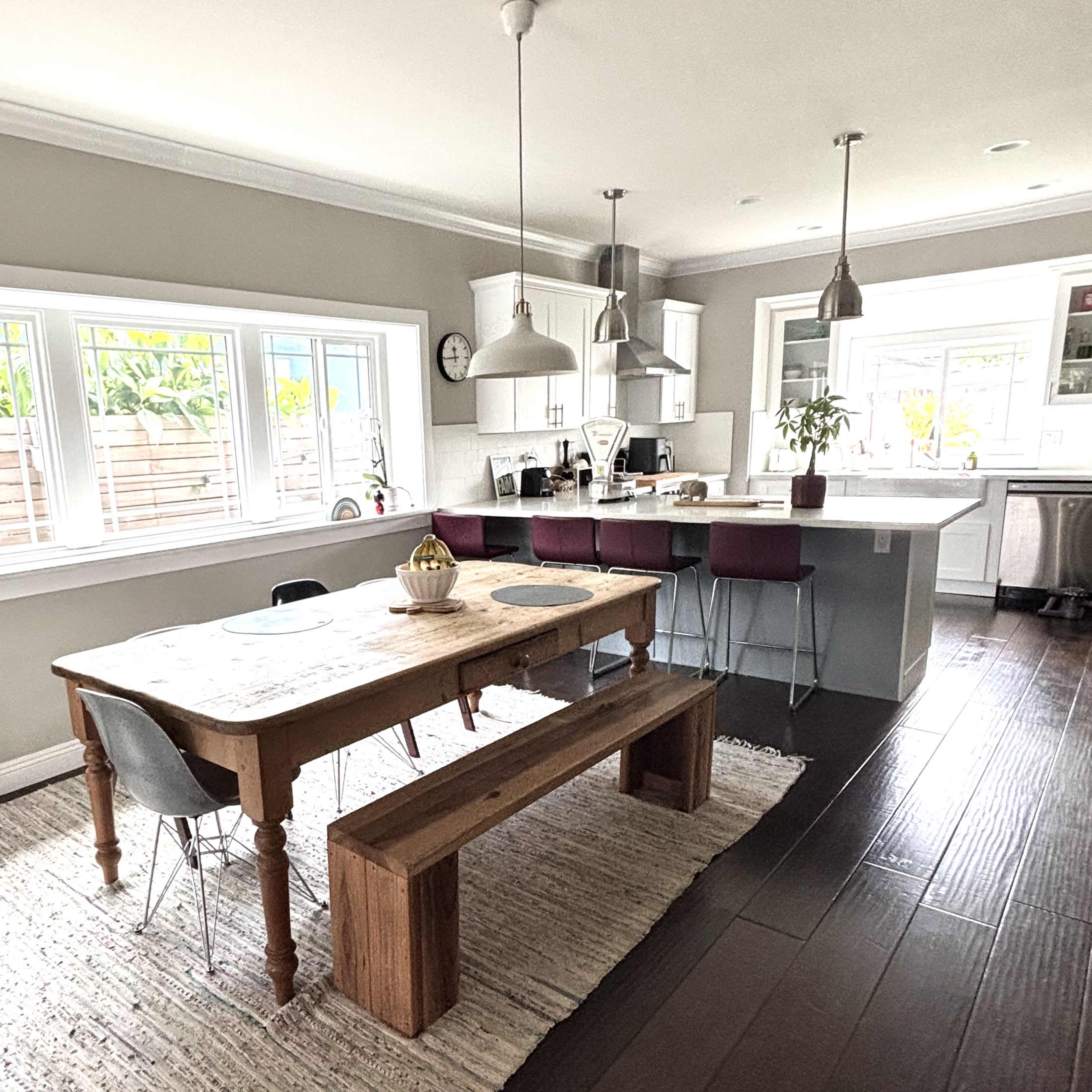 A modern, open-concept kitchen and dining area features a wooden dining table with a bench, surrounded by gray and purple chairs, with natural light streaming through large windows.