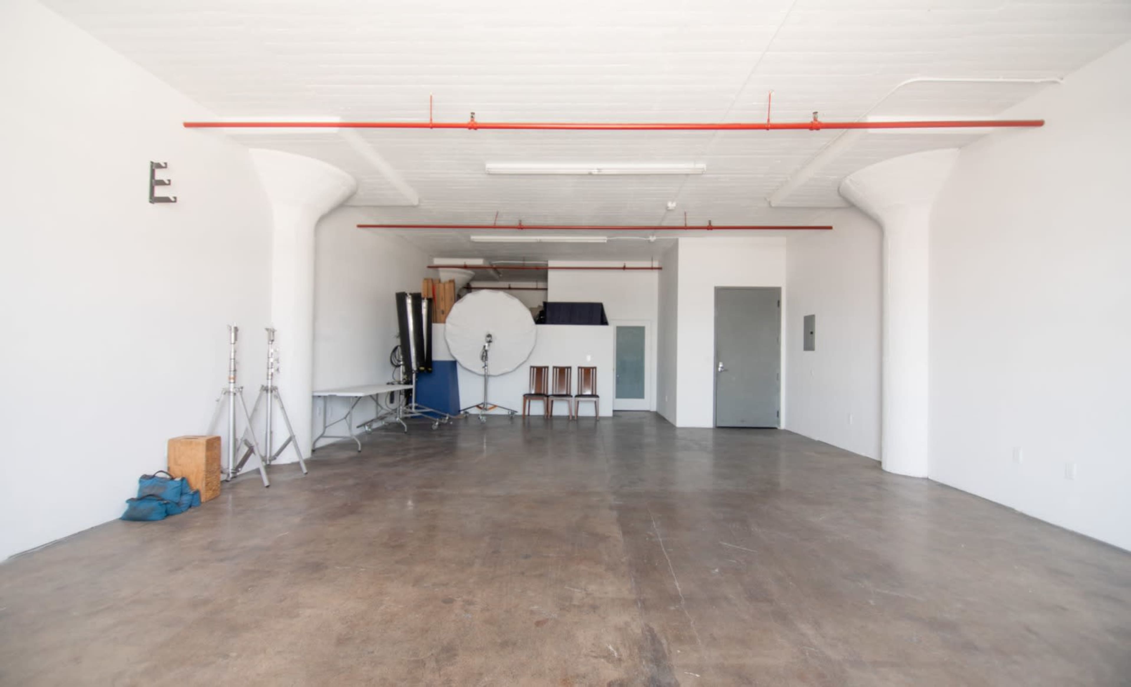 An empty studio space with concrete flooring, white walls, and various equipment including stands and a round backdrop stored in the corner.
