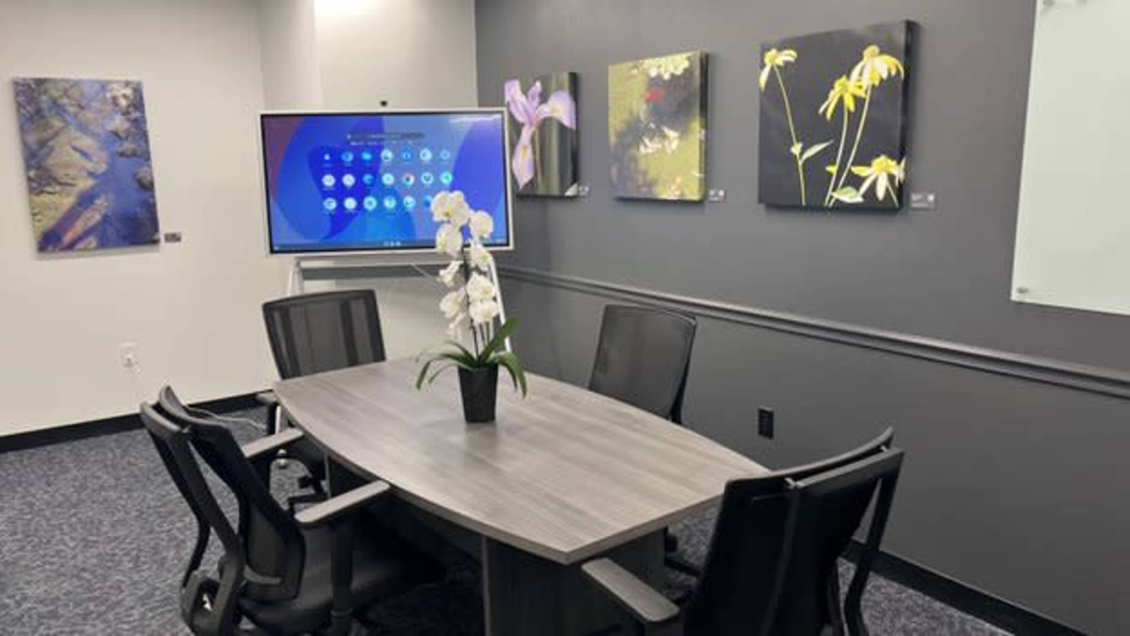 A conference room features a wooden table with four black chairs, a digital screen on the wall, and colorful floral artworks displayed.