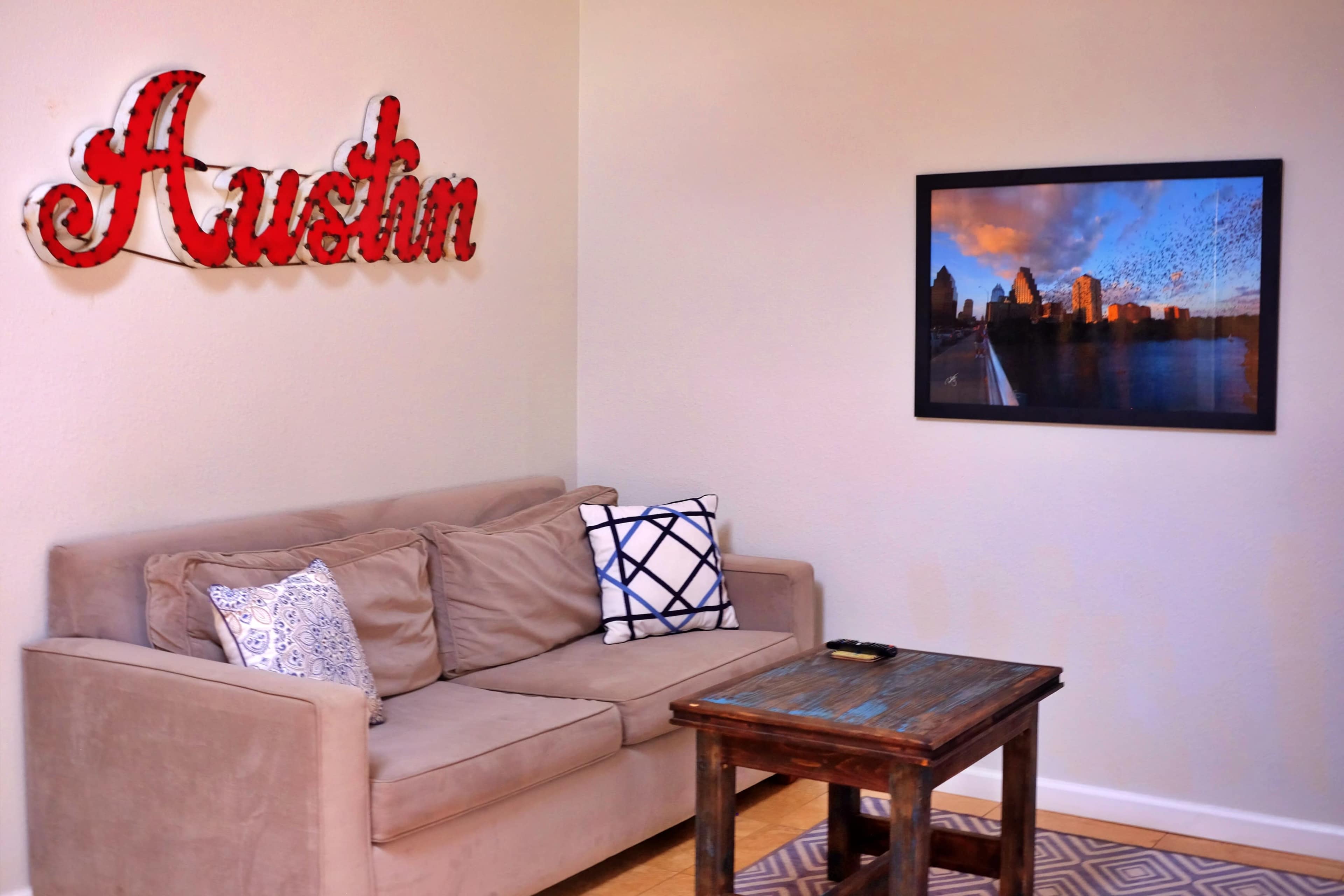 The image shows a living room corner with a beige sofa, a wooden coffee table, and a framed photograph of a city skyline on the wall.