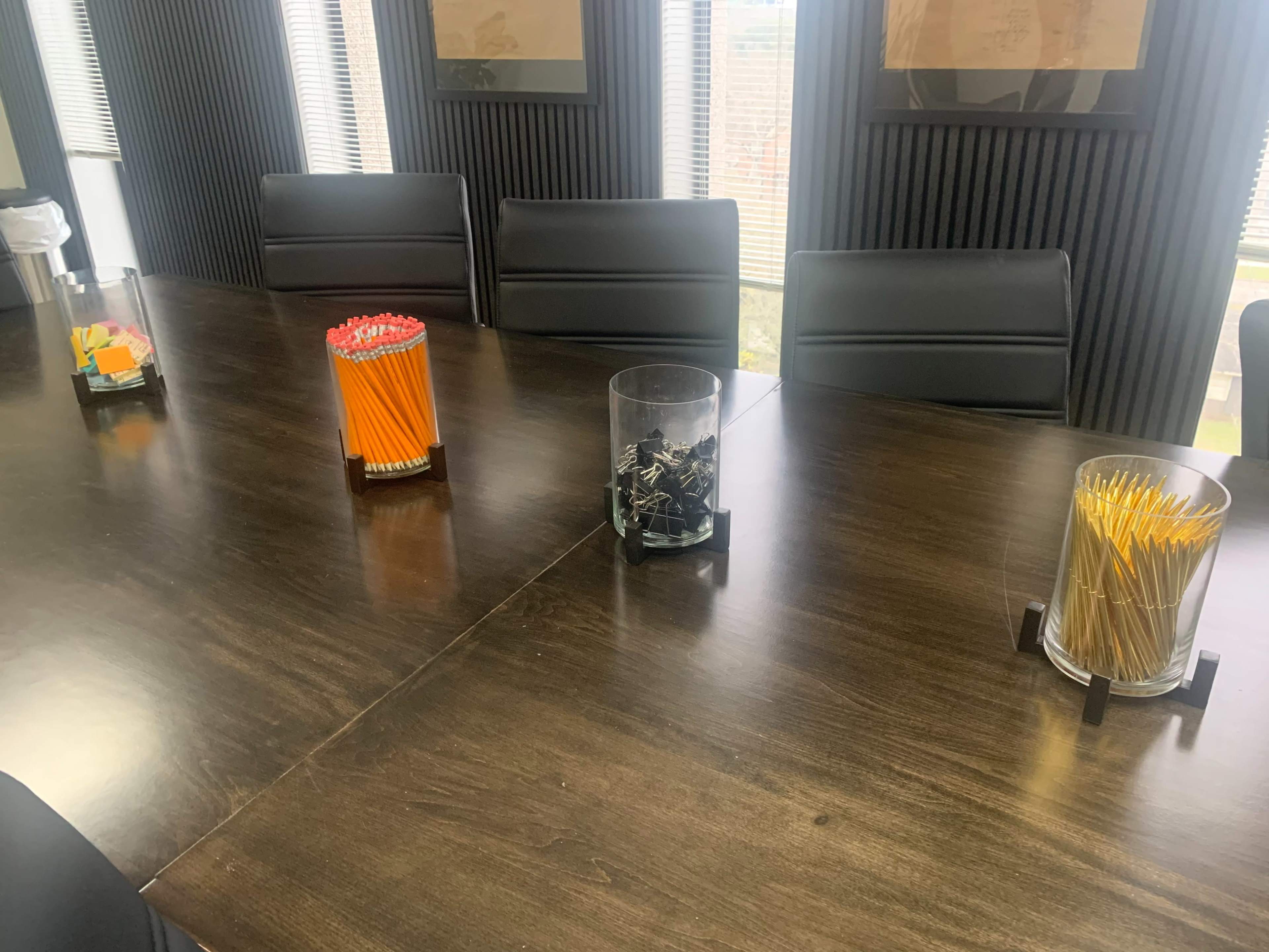A conference room table with several glass containers filled with office supplies, including pencils and paperclips, arranged neatly in front of empty chairs.
