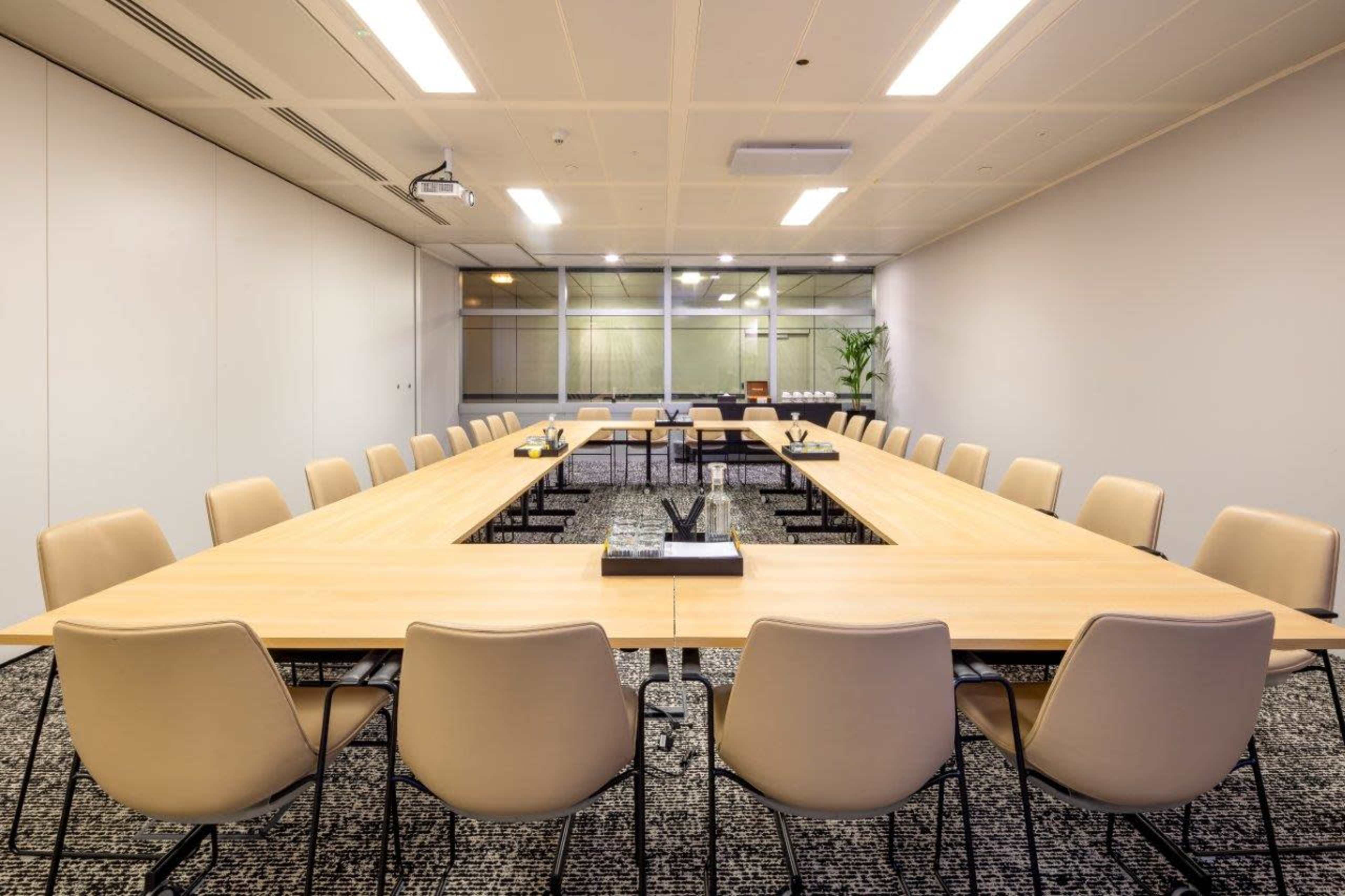 A large, rectangular conference room features a long wooden table surrounded by beige chairs, with a projector and glass windows in the background.