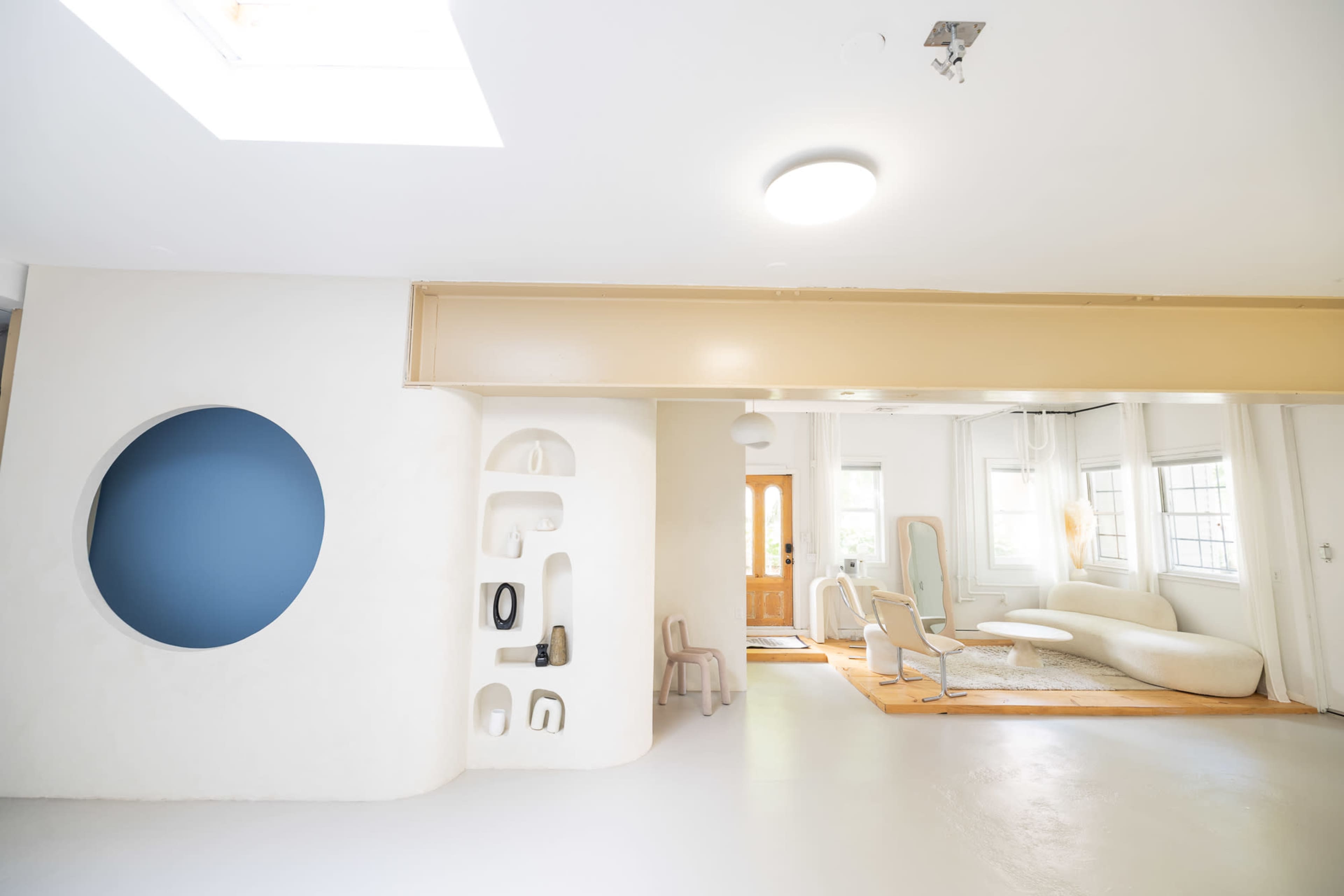 The image shows a modern, open-concept living space with a curved wall featuring a blue circular cutout, minimalistic furniture, and large windows allowing natural light.