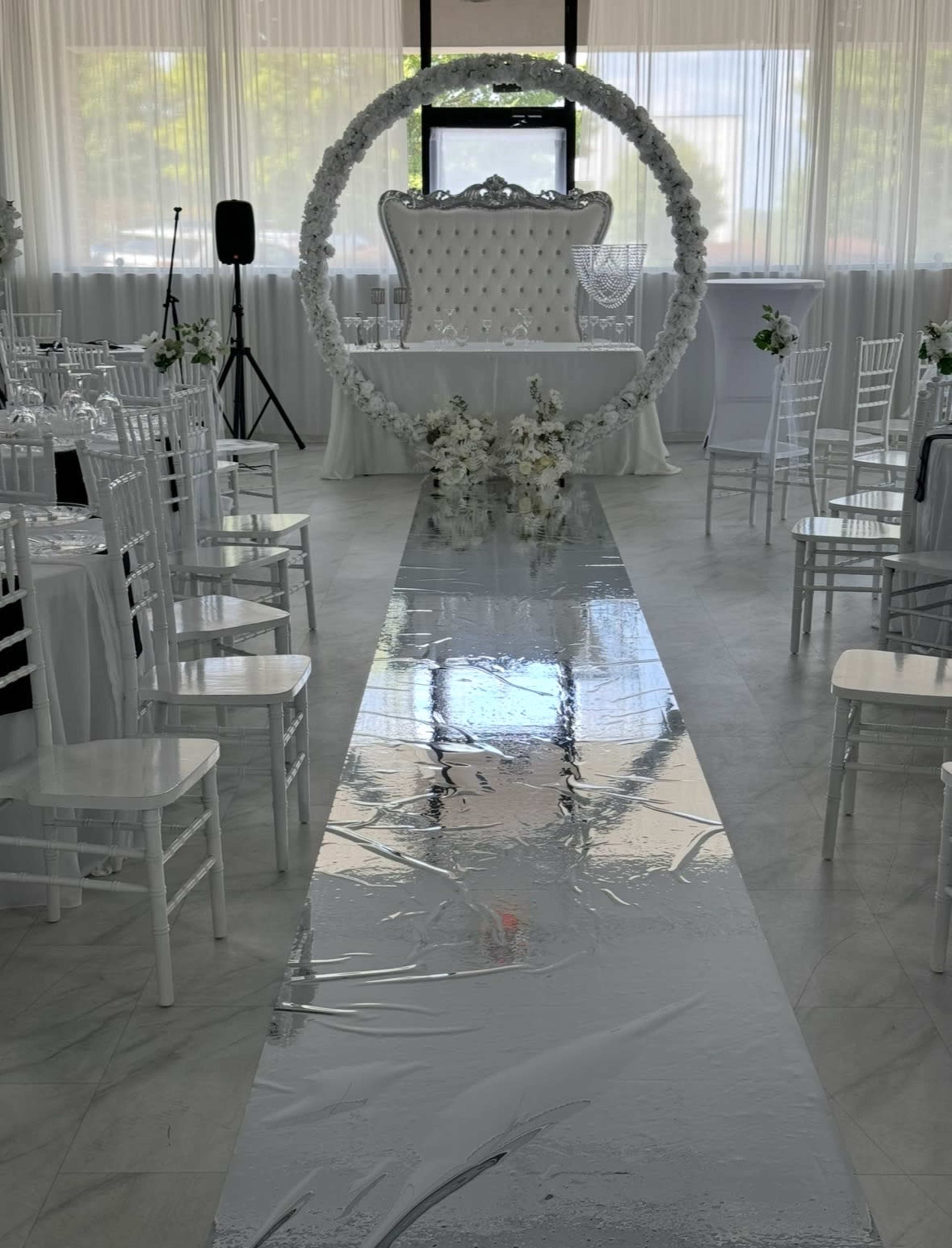 The image shows a wedding ceremony setup featuring a white aisle runner leading to a decorated arch and an elegant white throne at the front of a bright, airy space filled with white chairs.