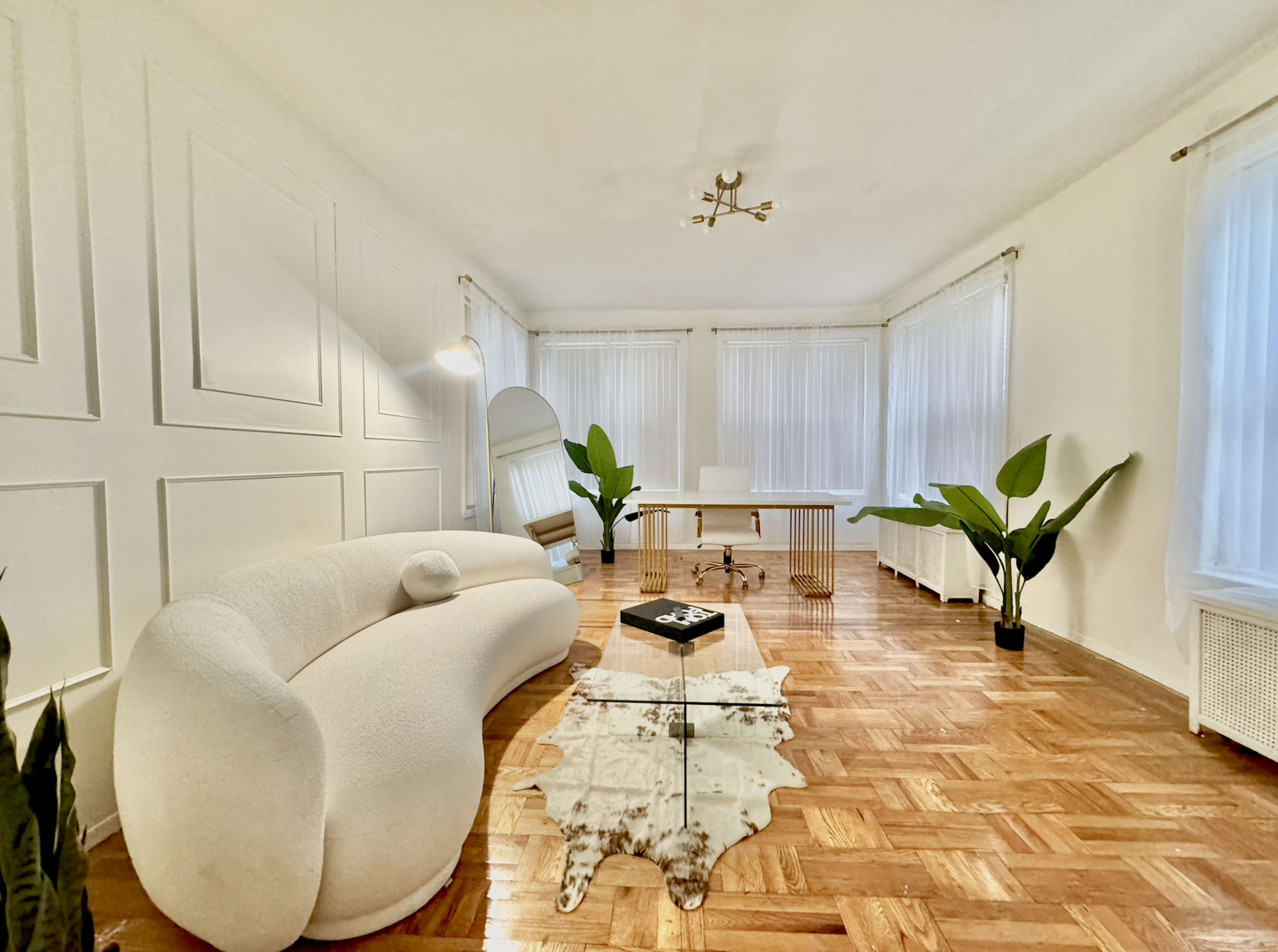 A spacious, well-lit room features a curved white sofa, a wooden desk, and decorative plants, all complemented by light-colored walls and large windows.