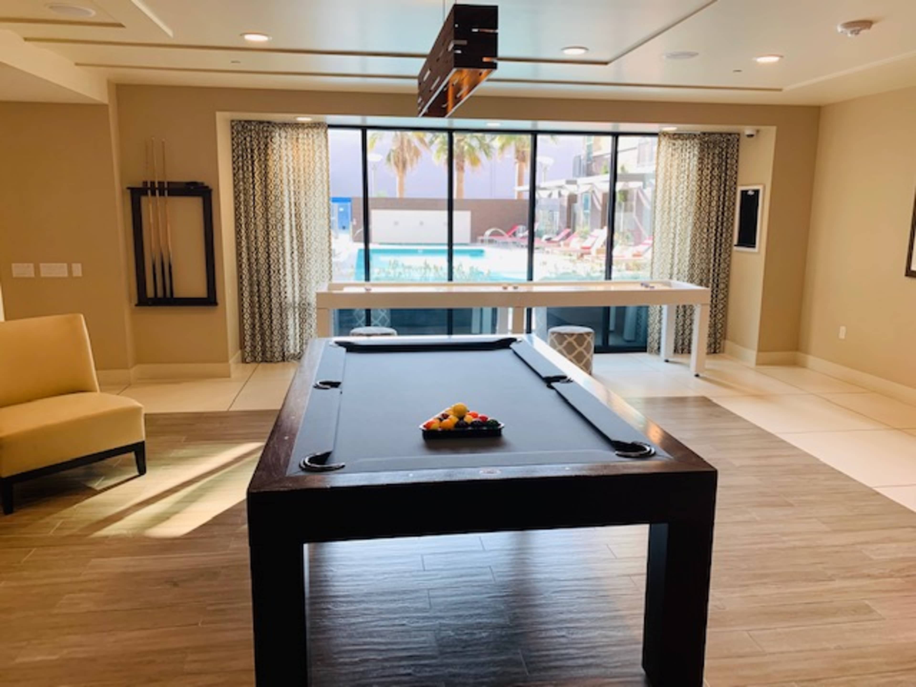 A well-lit room featuring a pool table, a window with a view of a swimming pool, and a seating area.