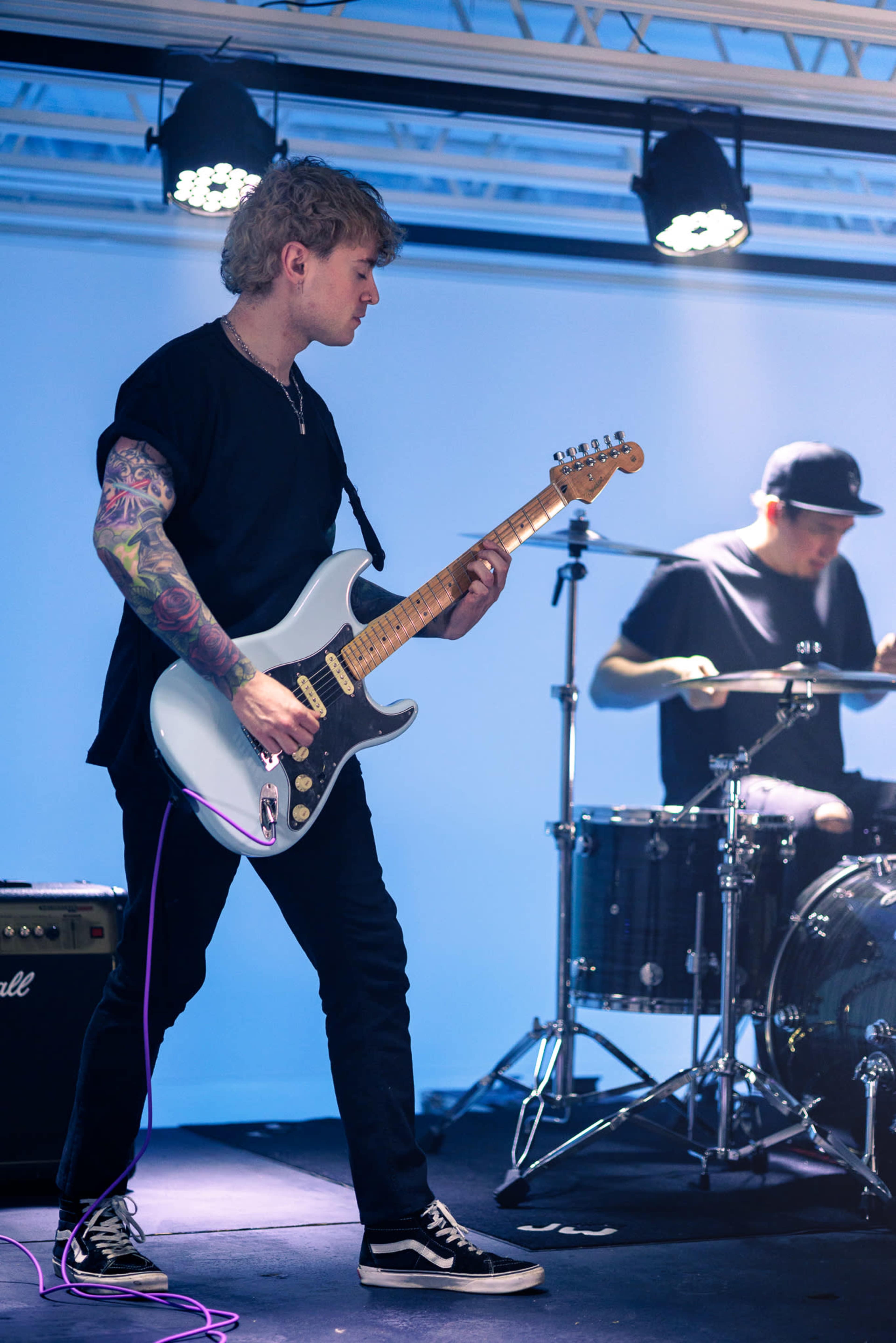 A guitarist performs on stage with a drummer in the background during a live music event.