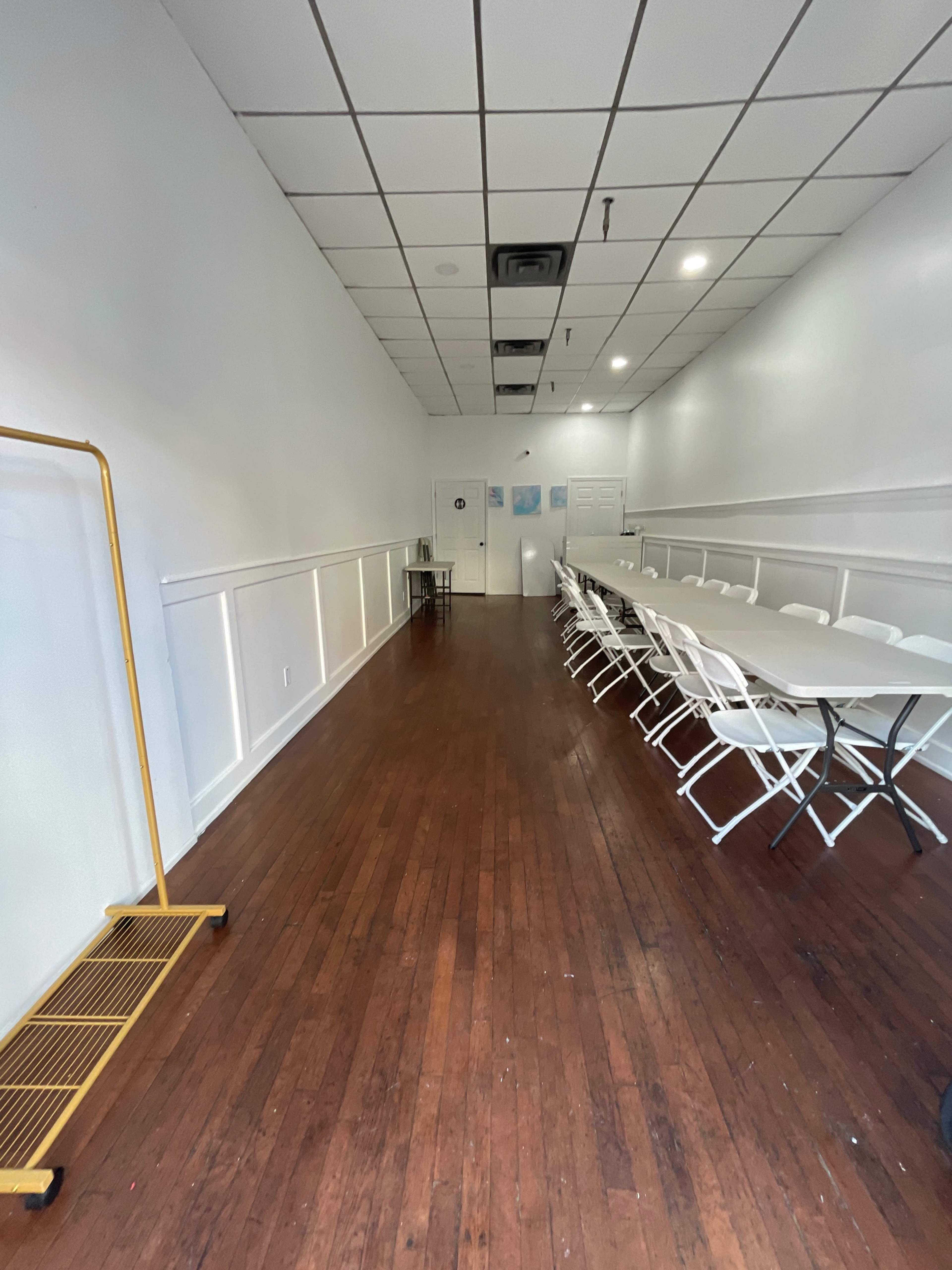 A long, narrow room with a wooden floor, features a row of white folding chairs and a long table, and has a rack along one side.