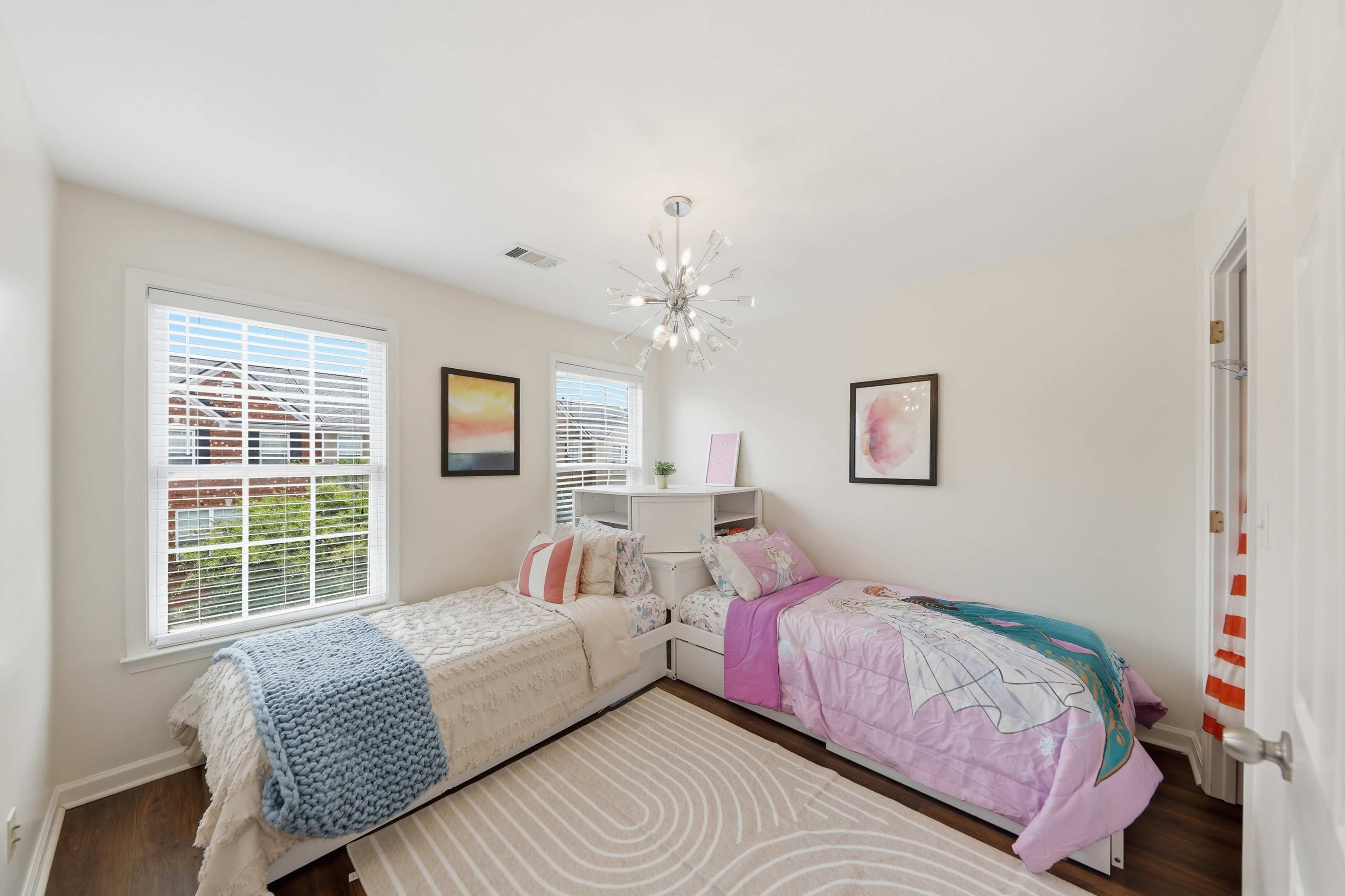 The image shows a bright bedroom with two beds, a light fixture, and large windows with natural light illuminating the space.