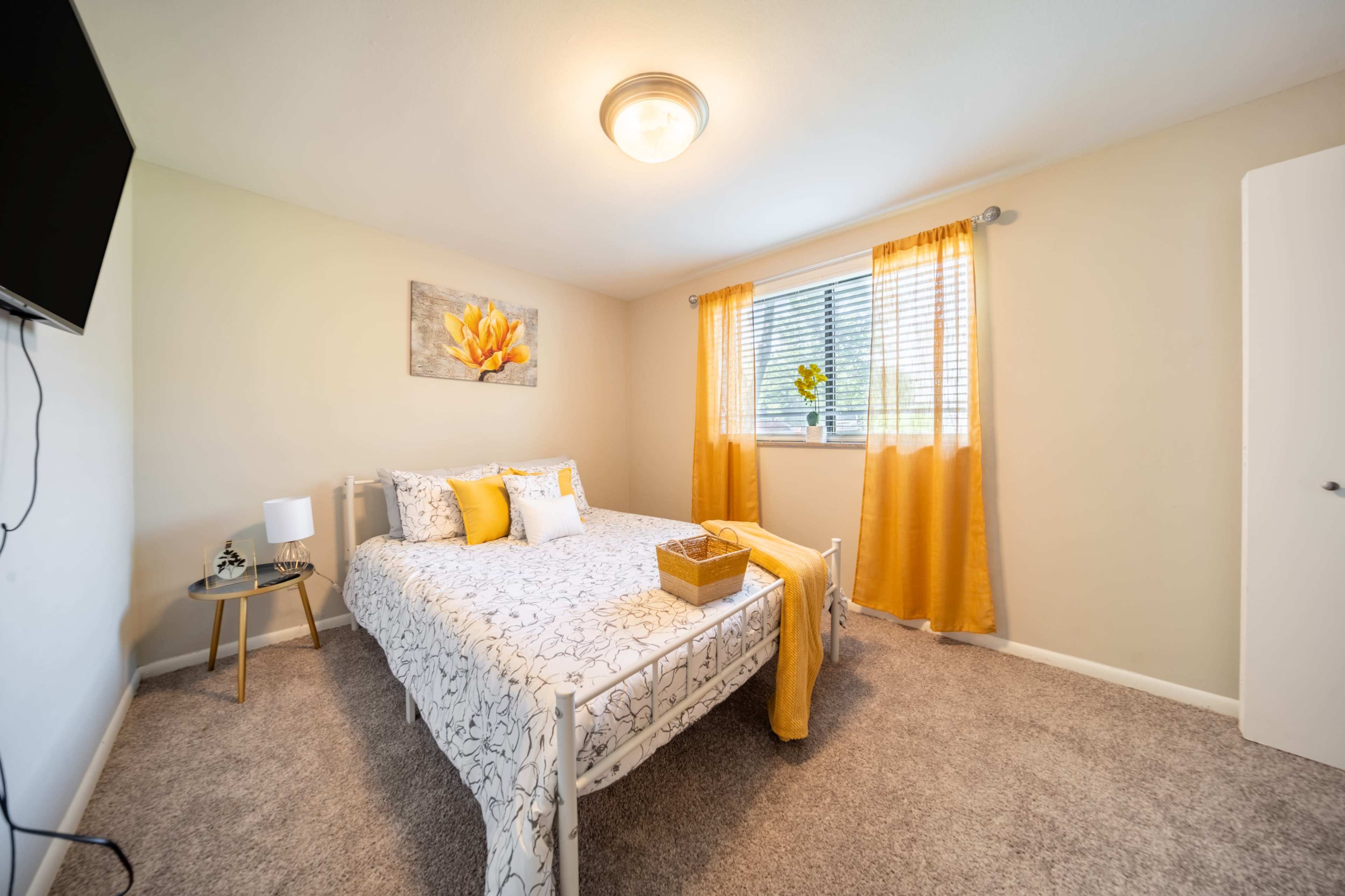 A neatly arranged bedroom with a bed, a nightstand, a wall-mounted TV, and large curtains beside a window.