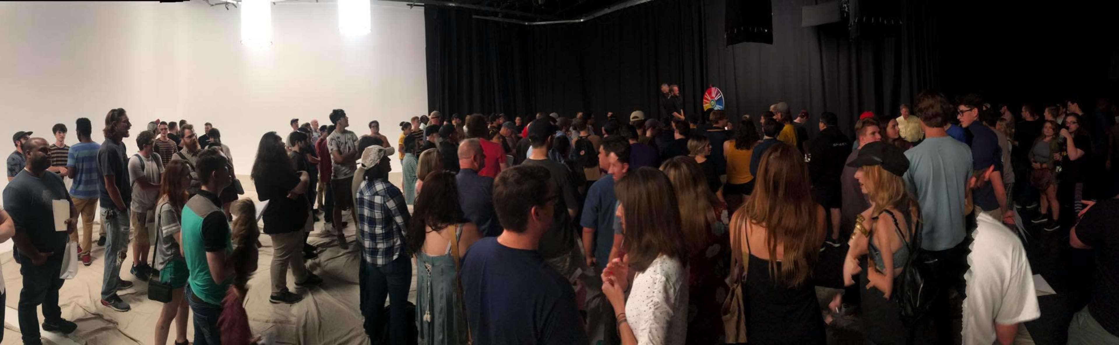 A large group of people is gathered in a dimly lit studio with a blank white backdrop for an event or performance.