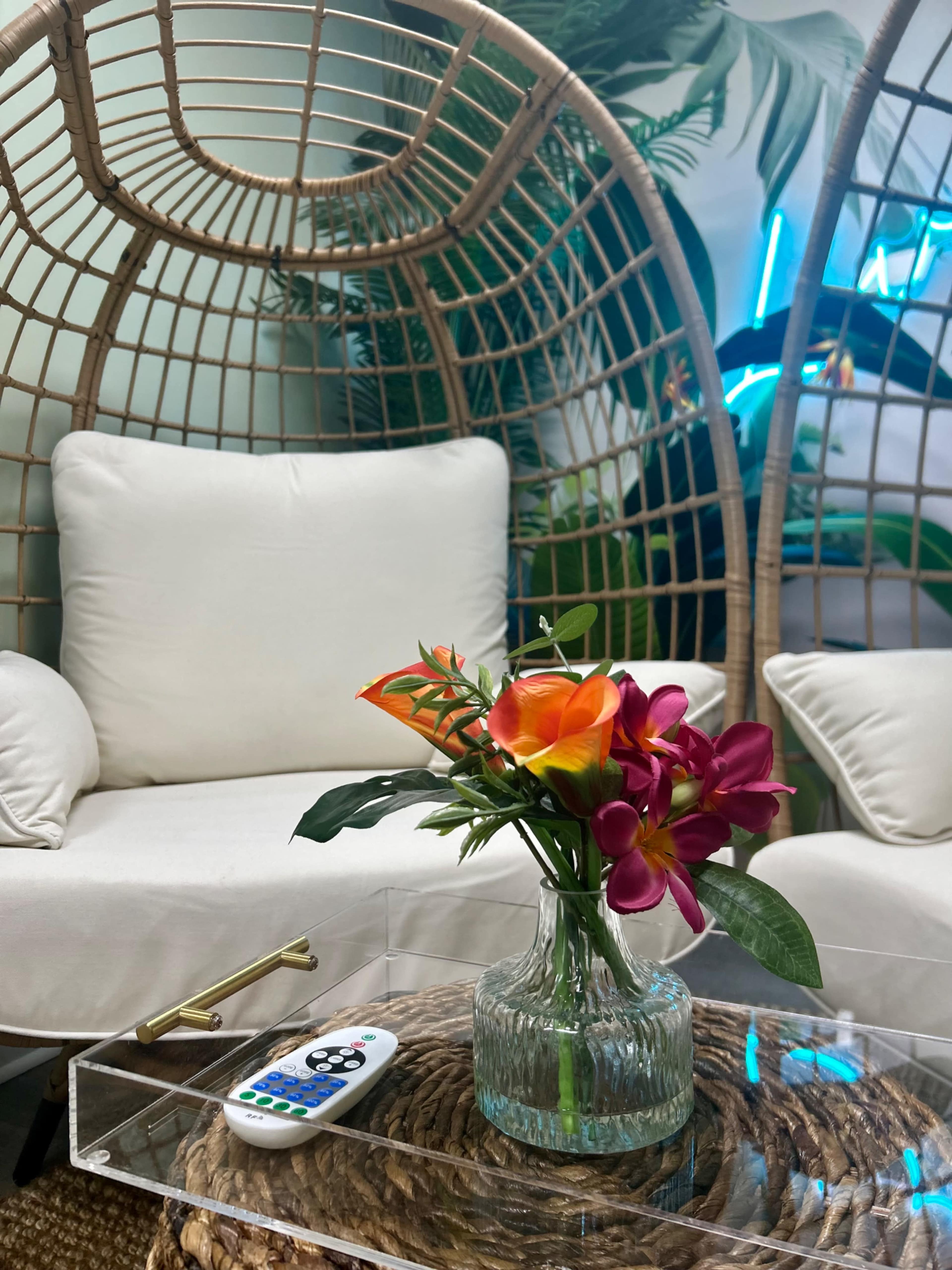 A glass vase with colorful flowers sits on a clear table next to a remote control, surrounded by two wicker chairs.
