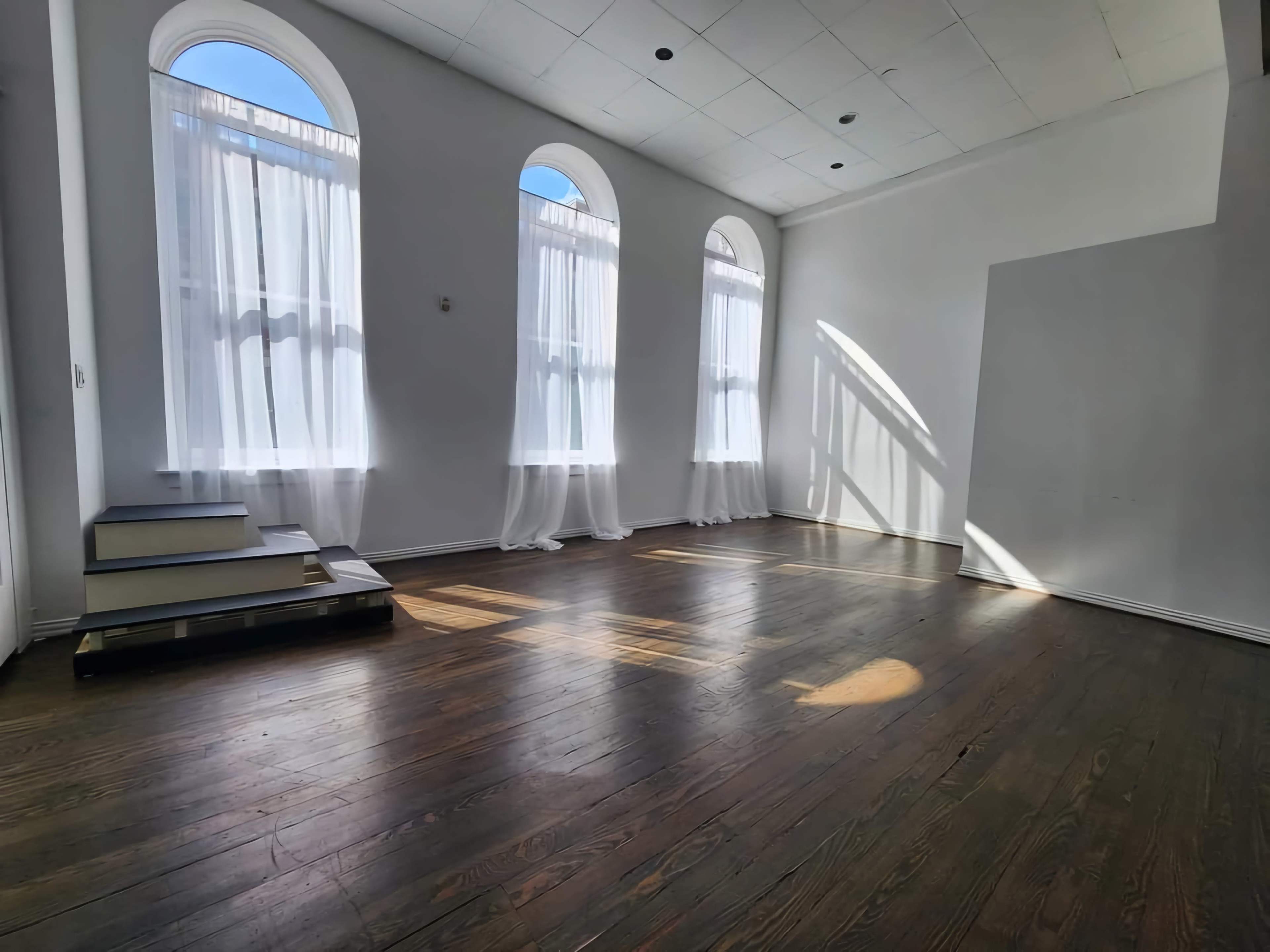 A bright room features large arched windows with sheer curtains, showcasing wooden flooring and a small set of steps.