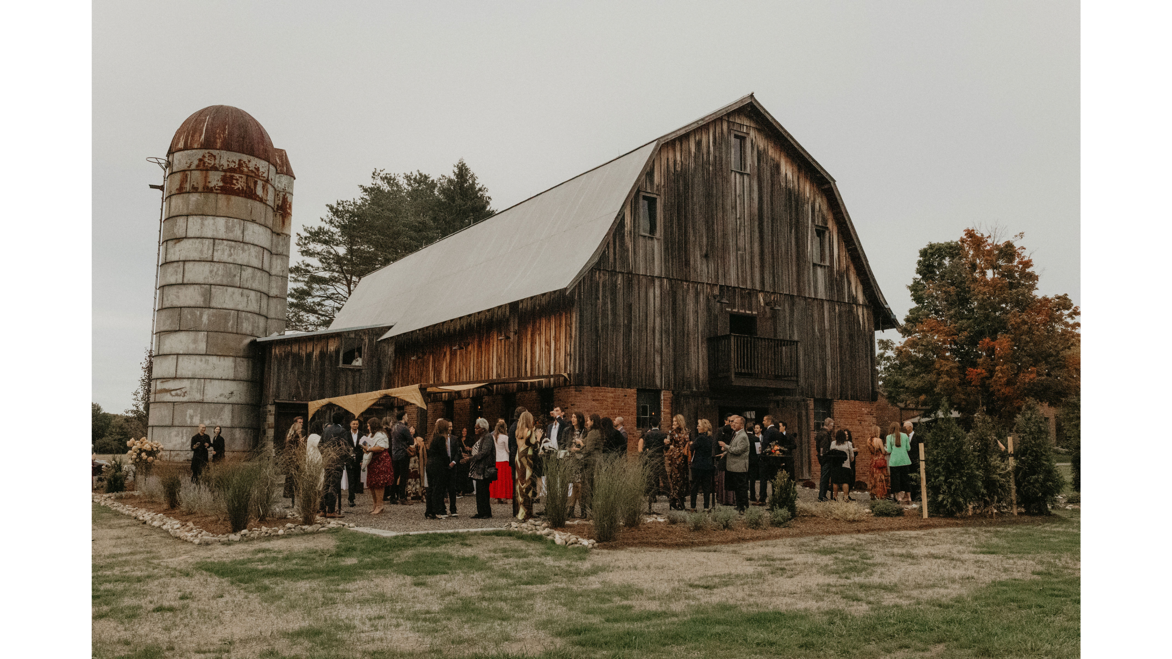 A gathering of people is taking place outside a rustic barn, with a silo nearby and trees in the background.