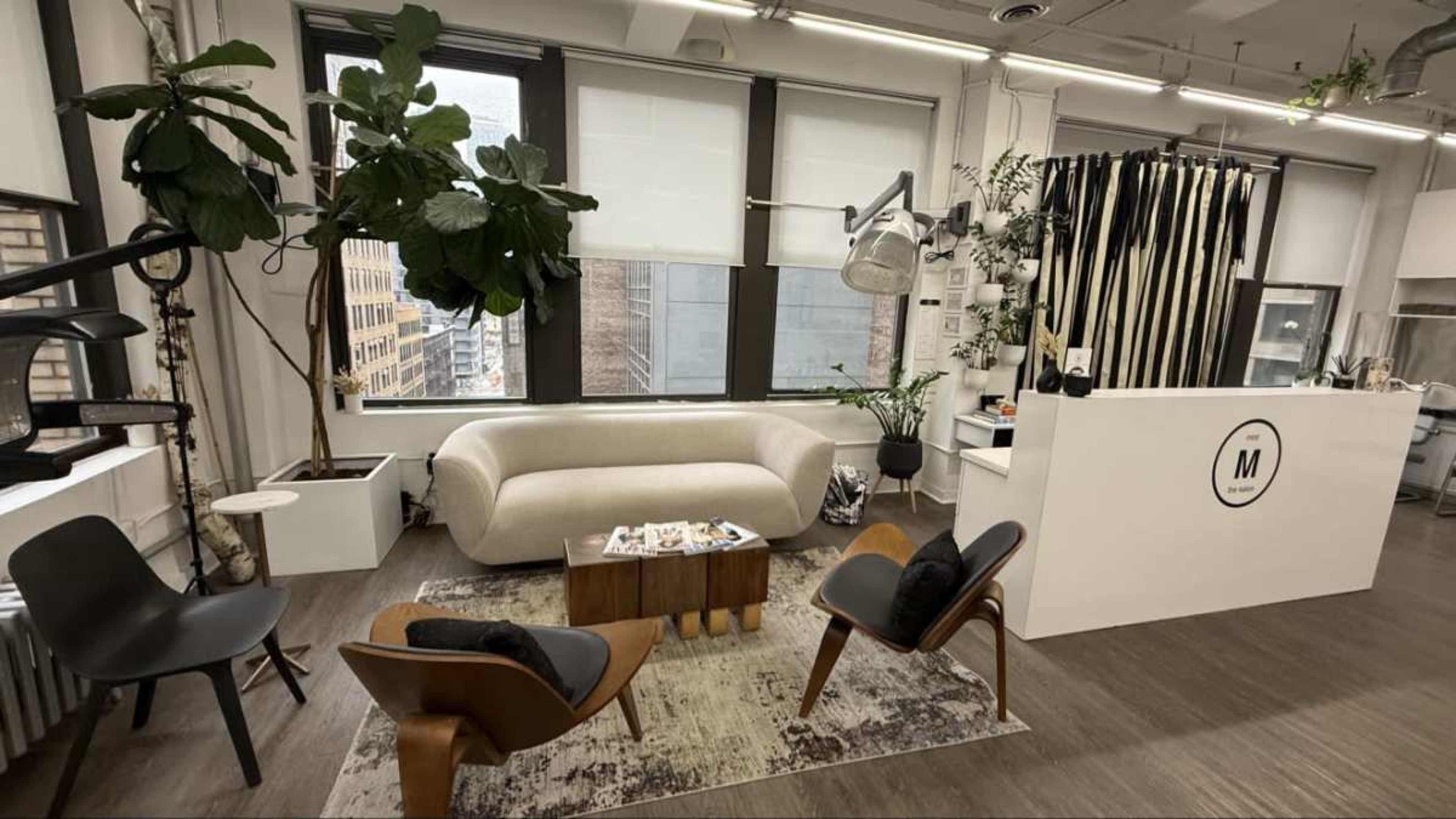 The image shows a stylish office space featuring a light-colored sofa, wooden chairs, a coffee table, and large windows with city views.