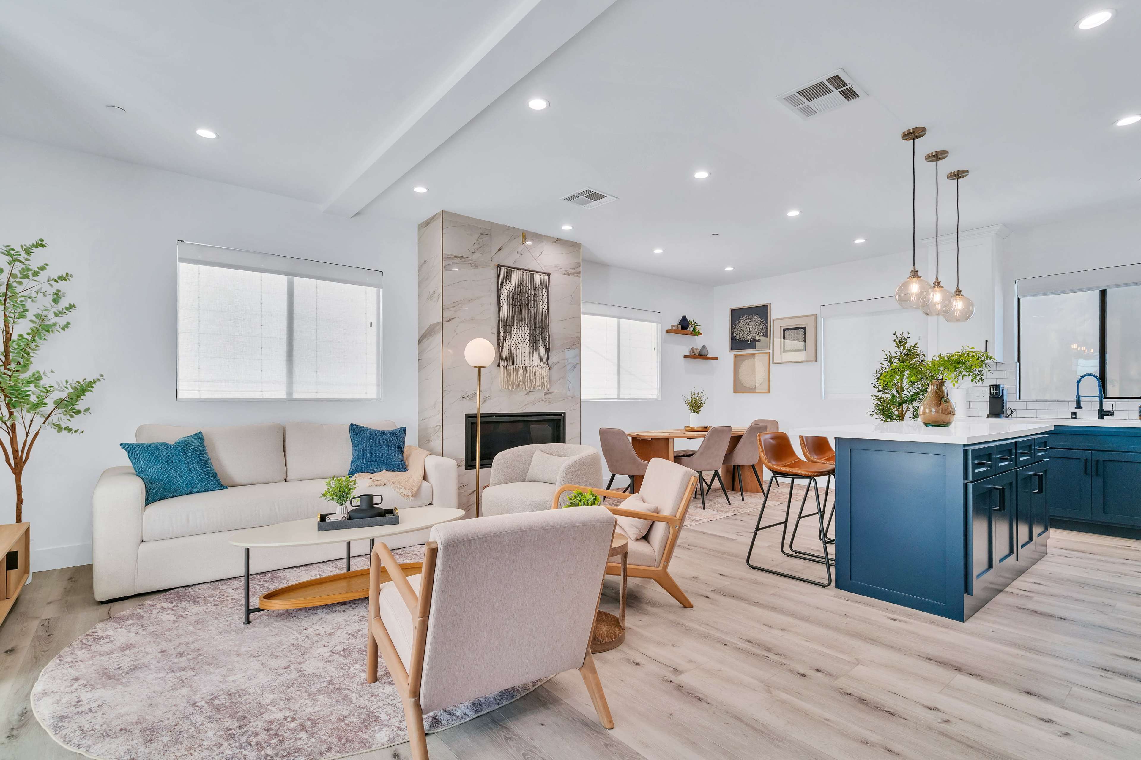 The image shows a modern living room and kitchen area with light-colored furniture, a central fireplace, and a dining table in the background.