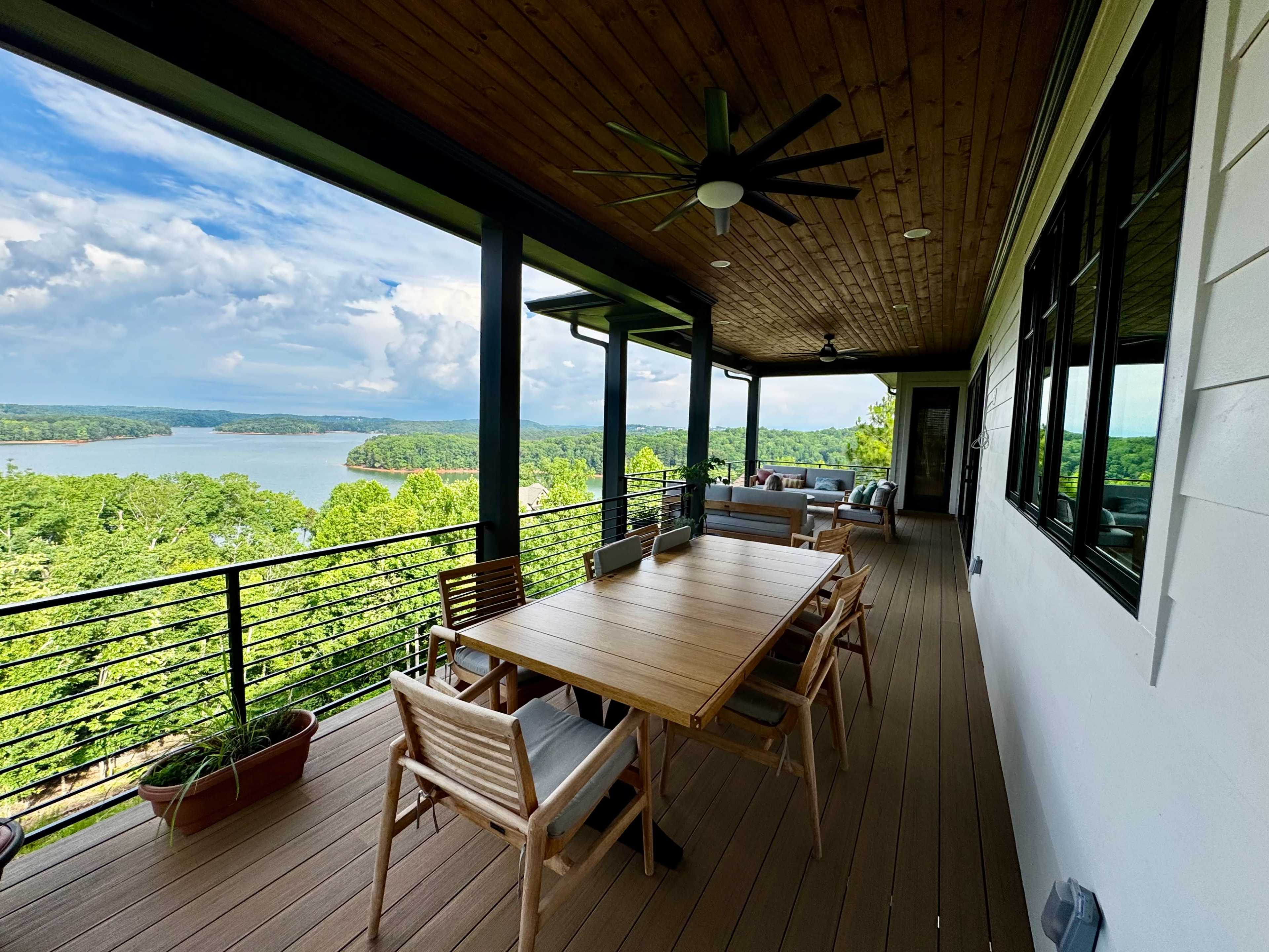 A covered deck features a large wooden table surrounded by chairs, overlooking a lake and lush greenery in the background.