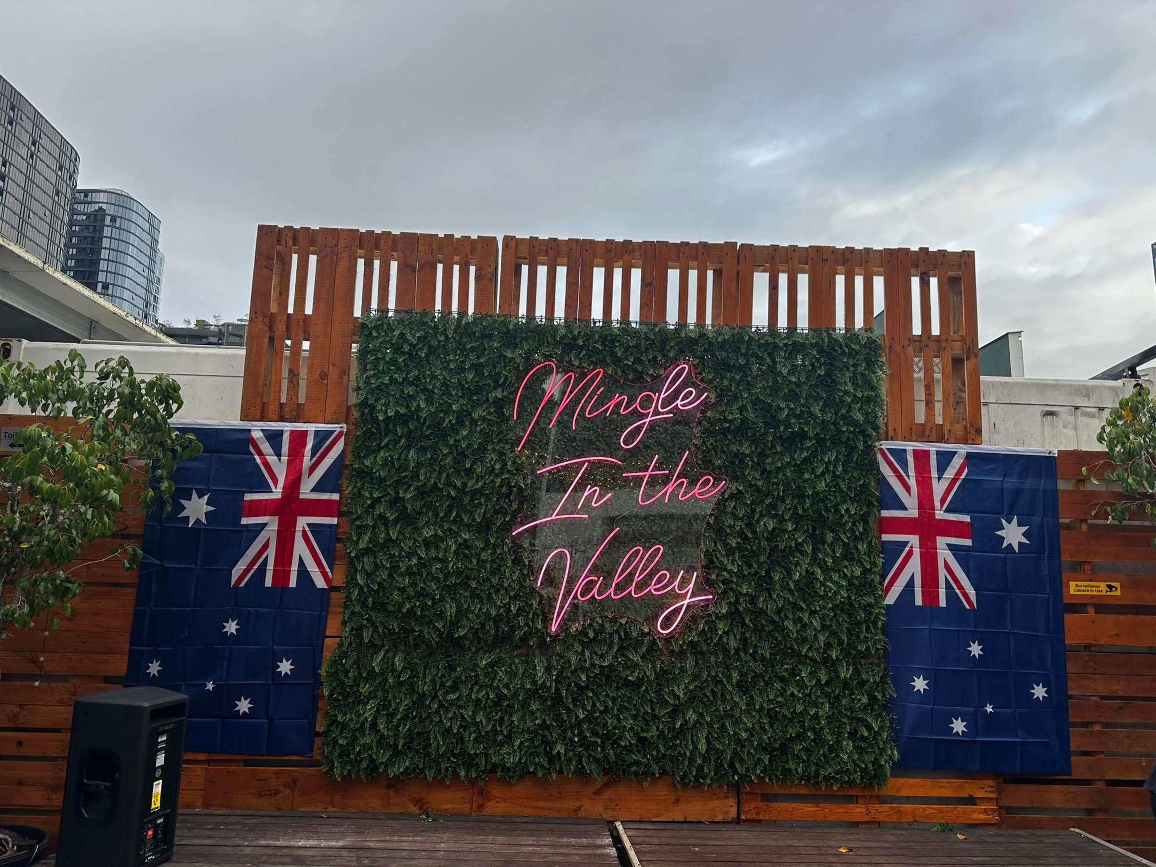 The image shows a vibrant outdoor space with a green foliage backdrop, featuring a neon sign that reads "Mingles In the Valley" flanked by Australian flags on either side.