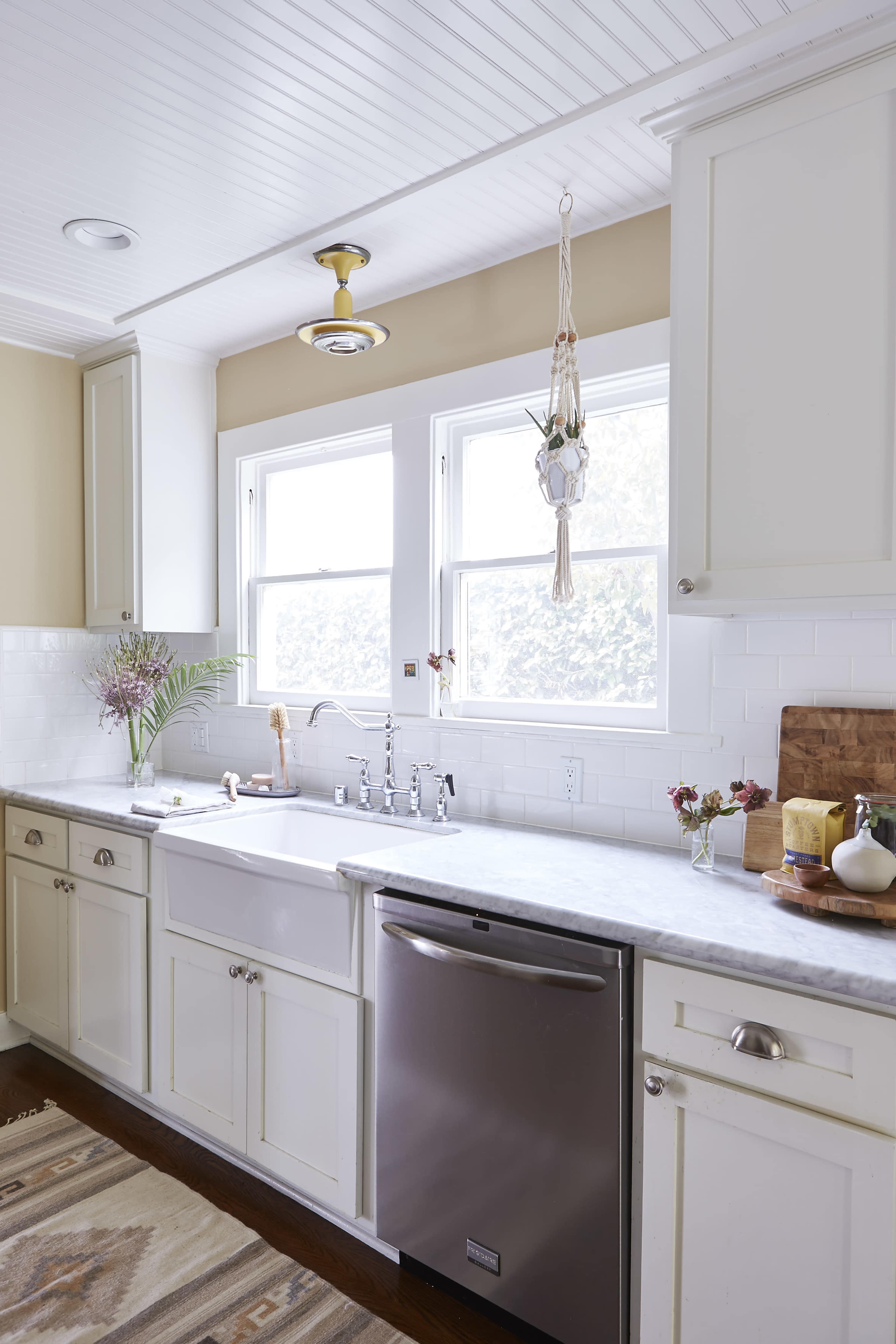A bright kitchen featuring a white farm sink, stainless steel dishwasher, and a marble countertop, with hanging plants near the window.