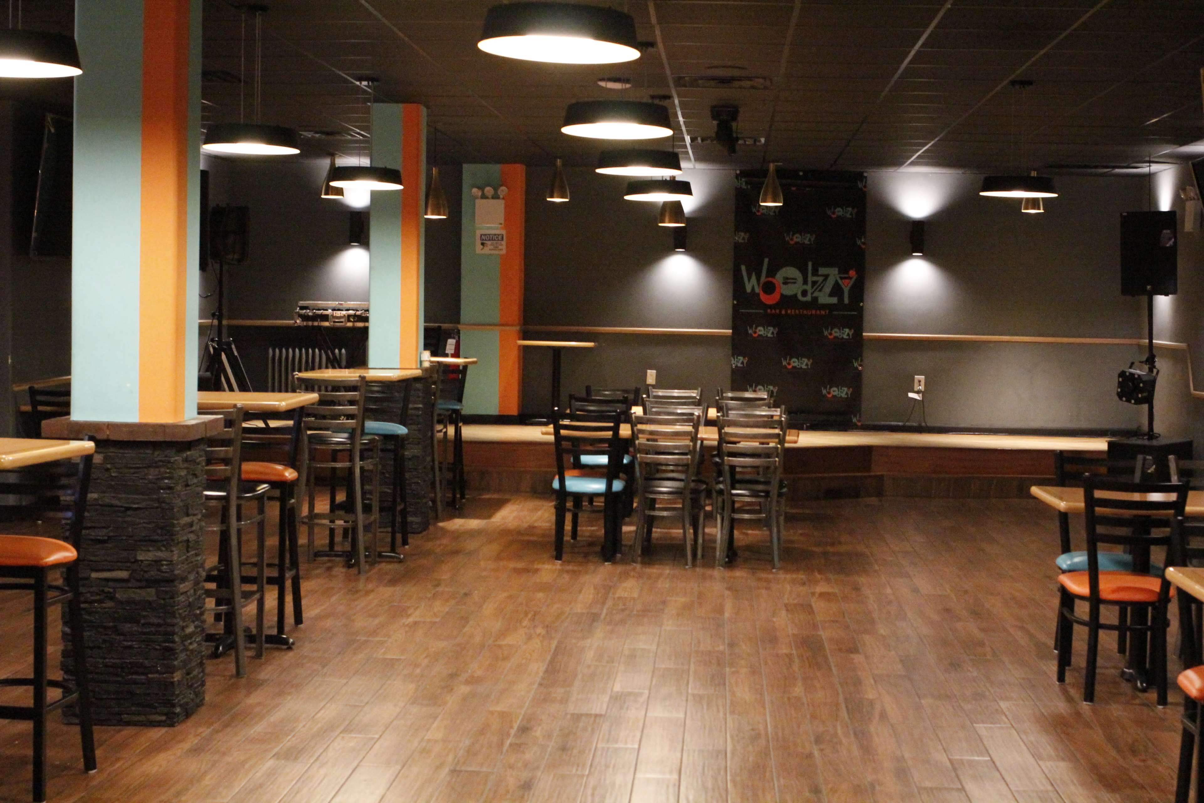 An empty bar area with wooden floors, several tables, and chairs arranged in a spacious layout, along with a small stage and sound equipment in the background.