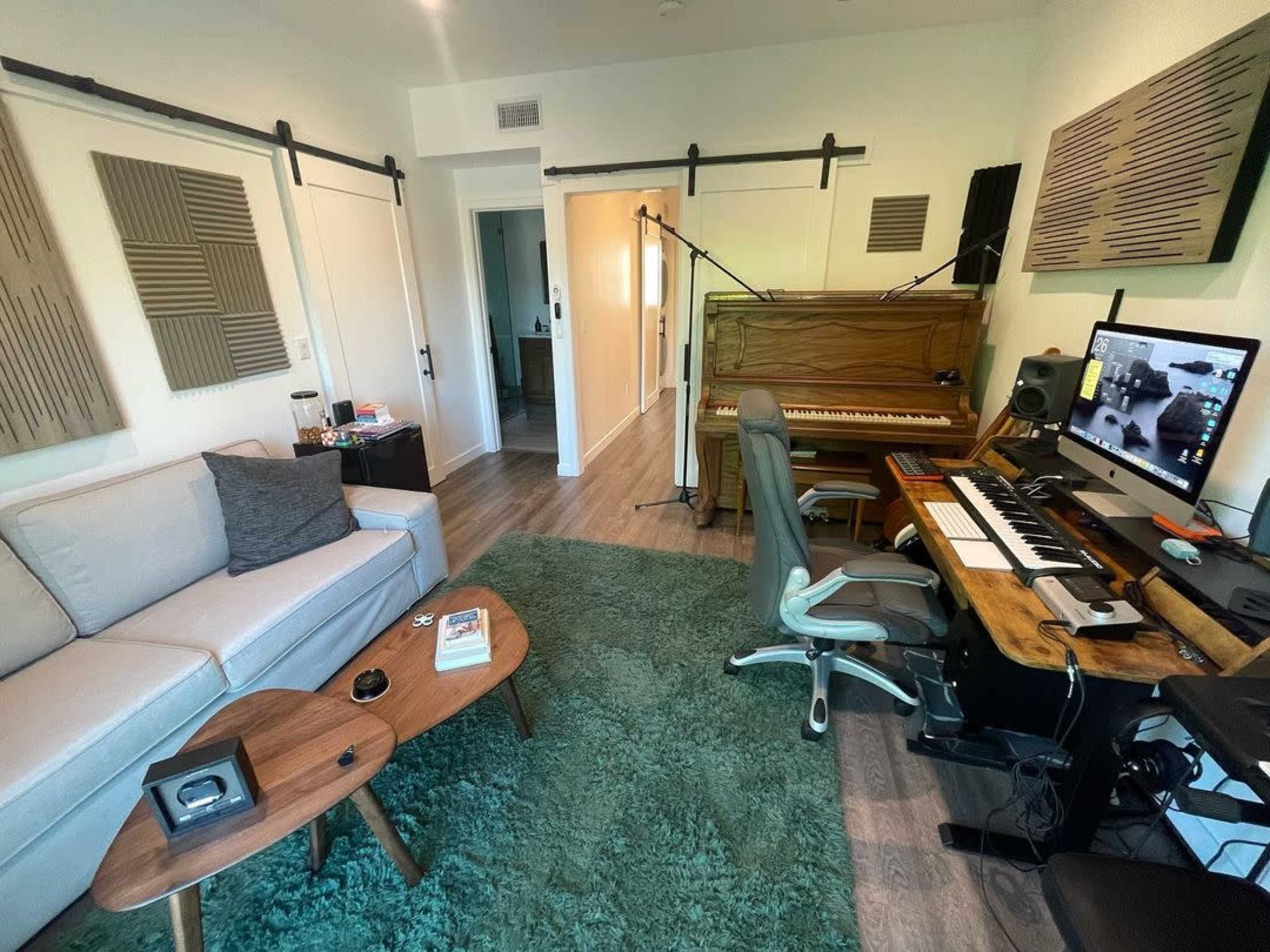 The image shows a cozy music studio featuring a piano, a computer setup with a keyboard, a sofa, and acoustic panels on the walls.