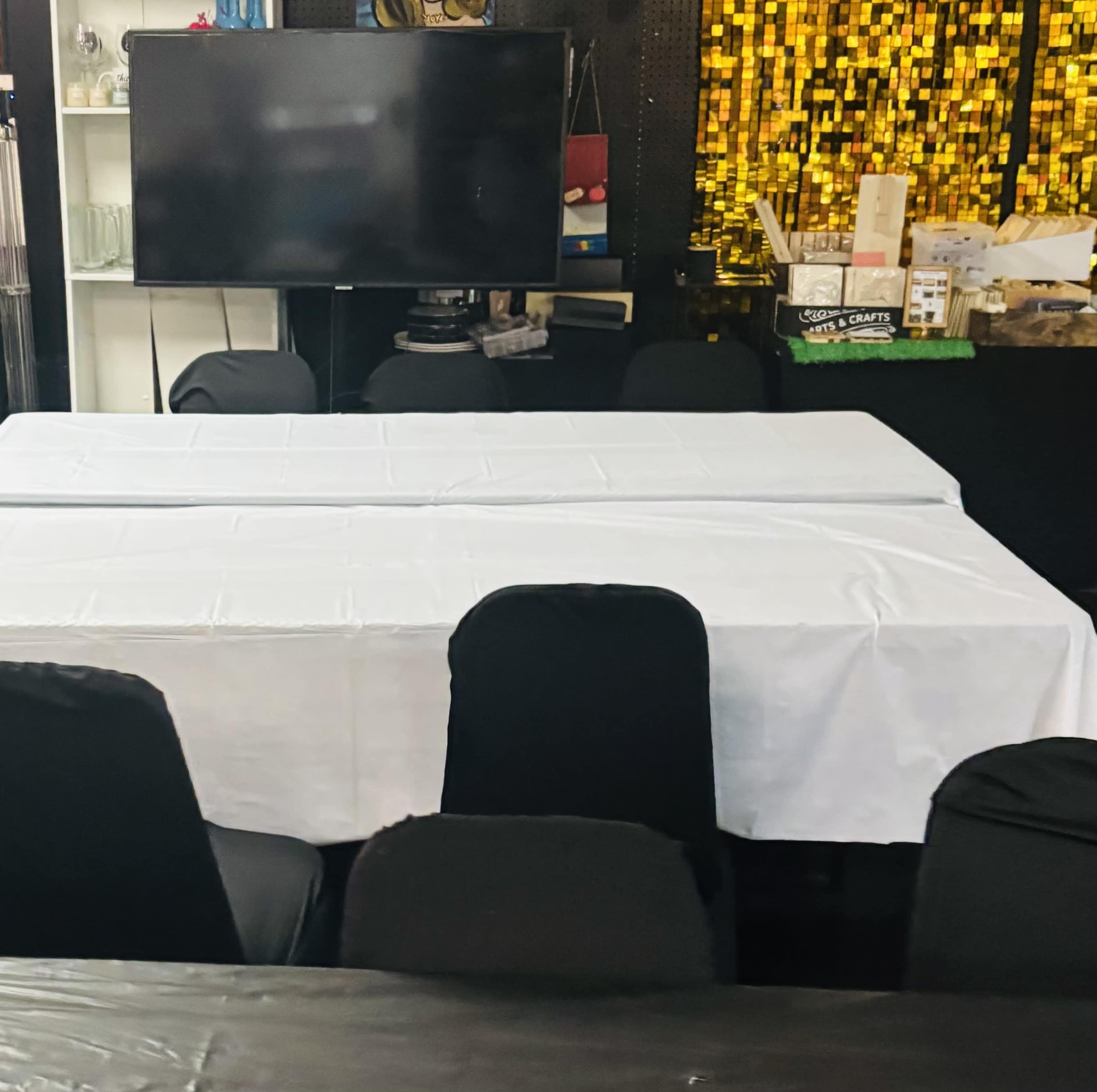 The image shows a set of tables covered with white tablecloths in a room with black chairs and a television in the background.