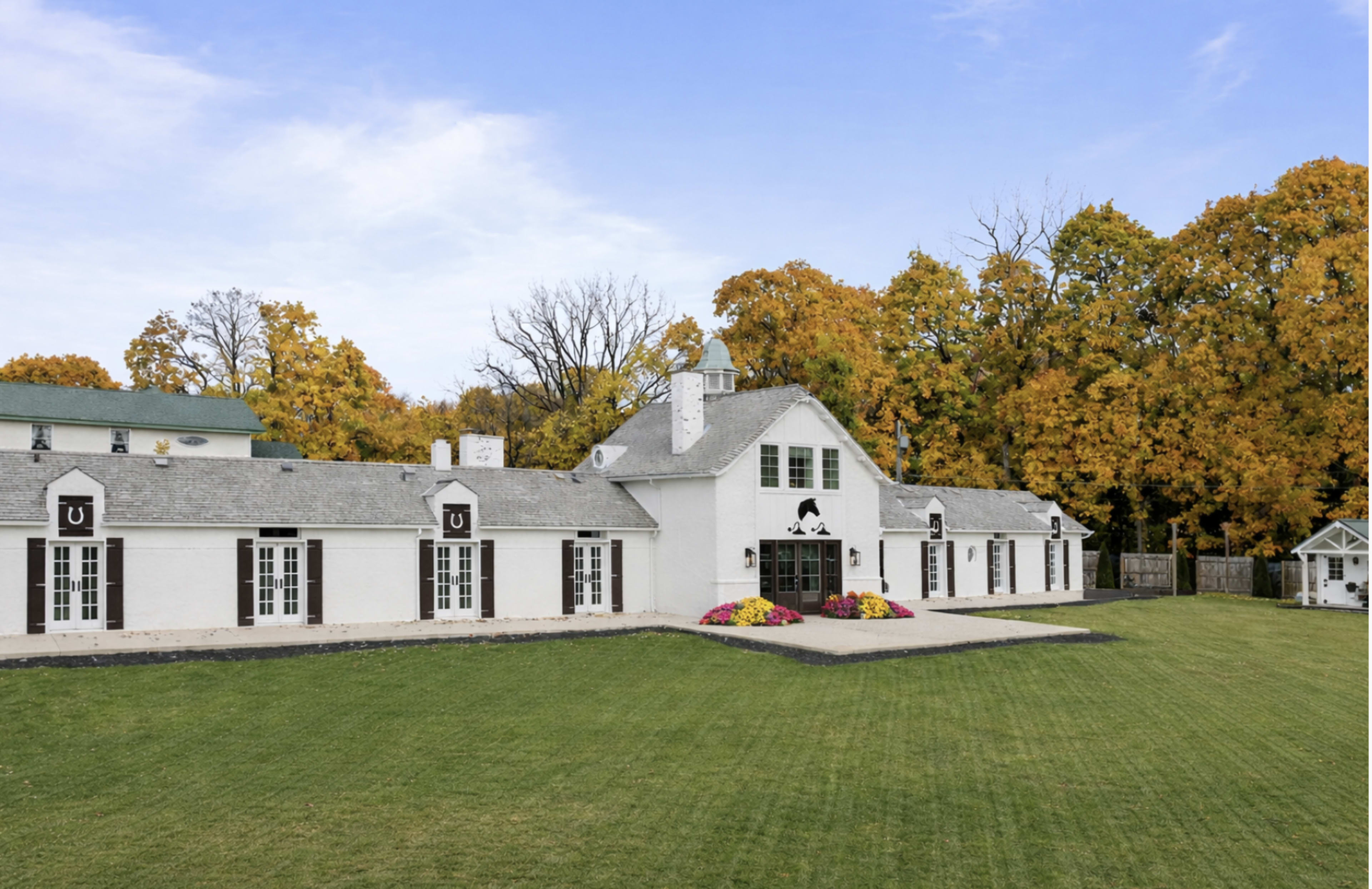 The image shows a spacious white building with a gray roof set against a backdrop of trees with autumn leaves.