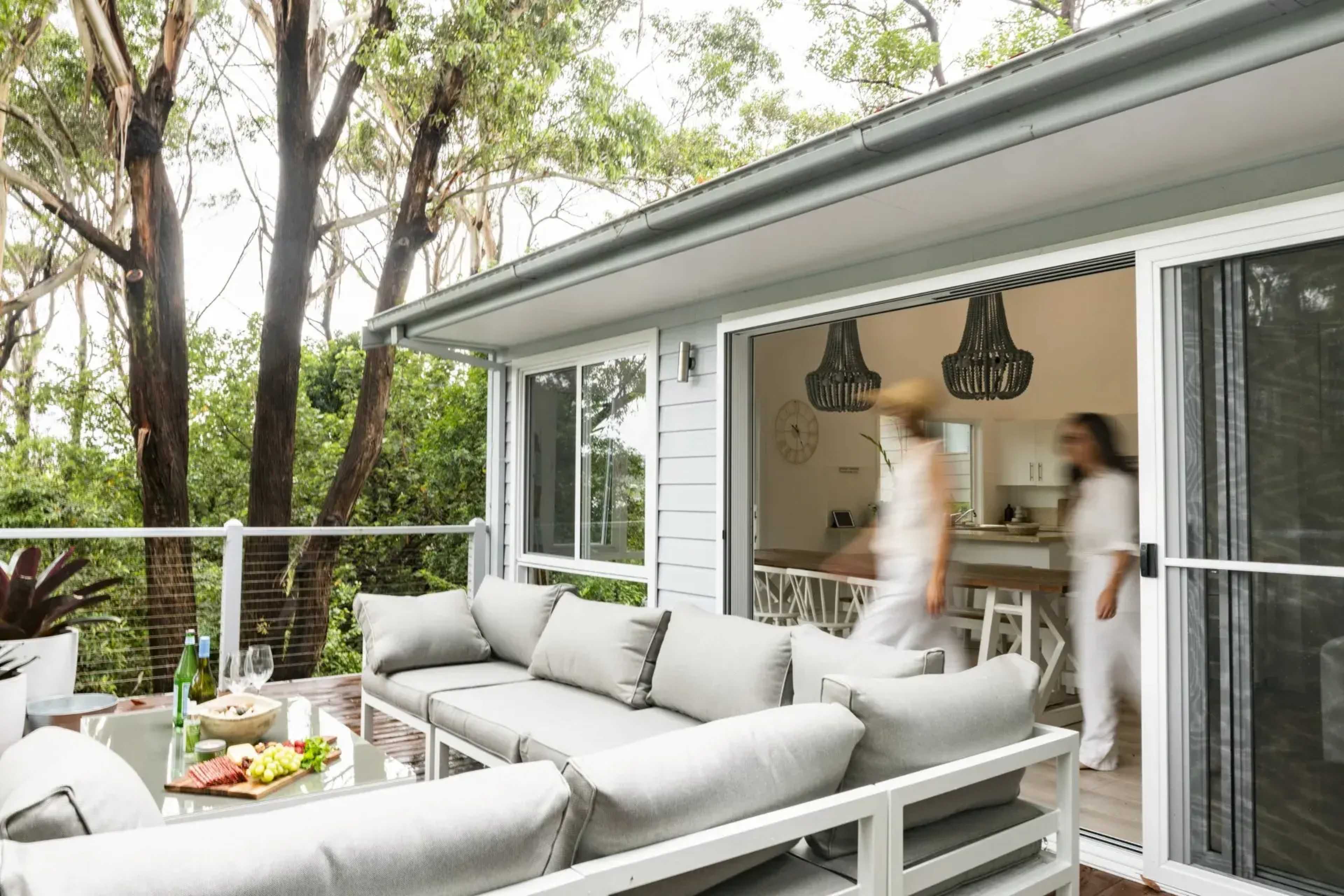 A person walks through an open doorway from a deck area with a large gray sofa into a kitchen and dining space surrounded by trees.