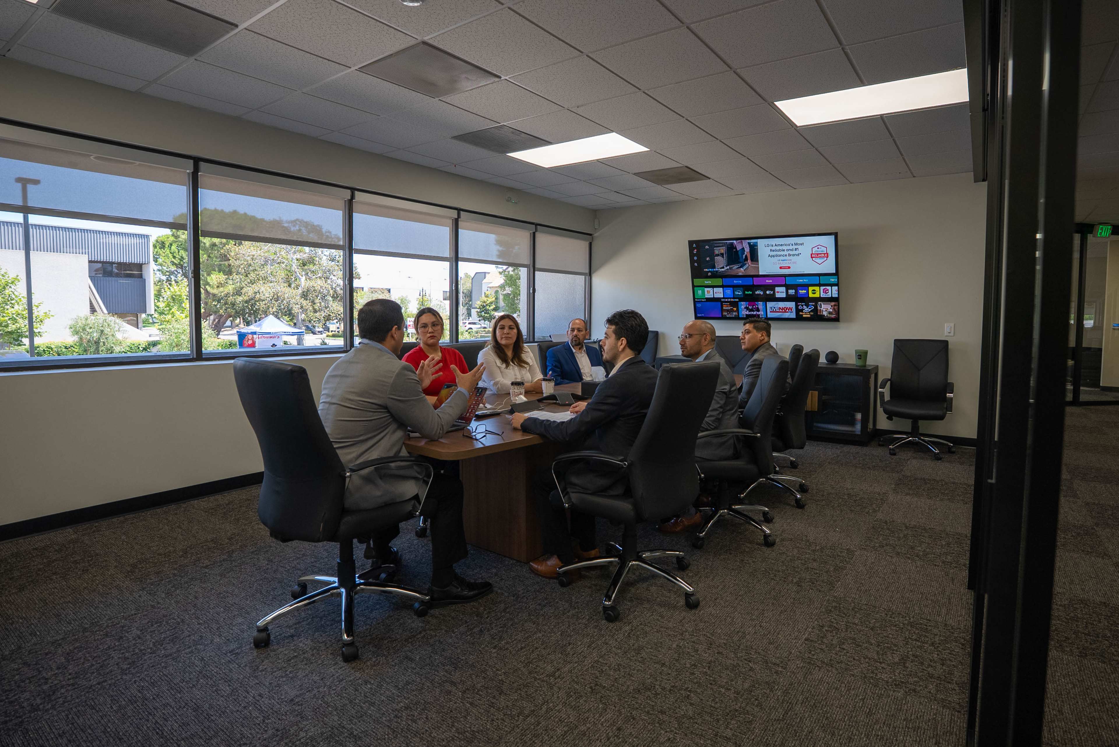 A group of six professionals is engaged in a meeting around a rectangular conference table in a well-lit office with large windows.