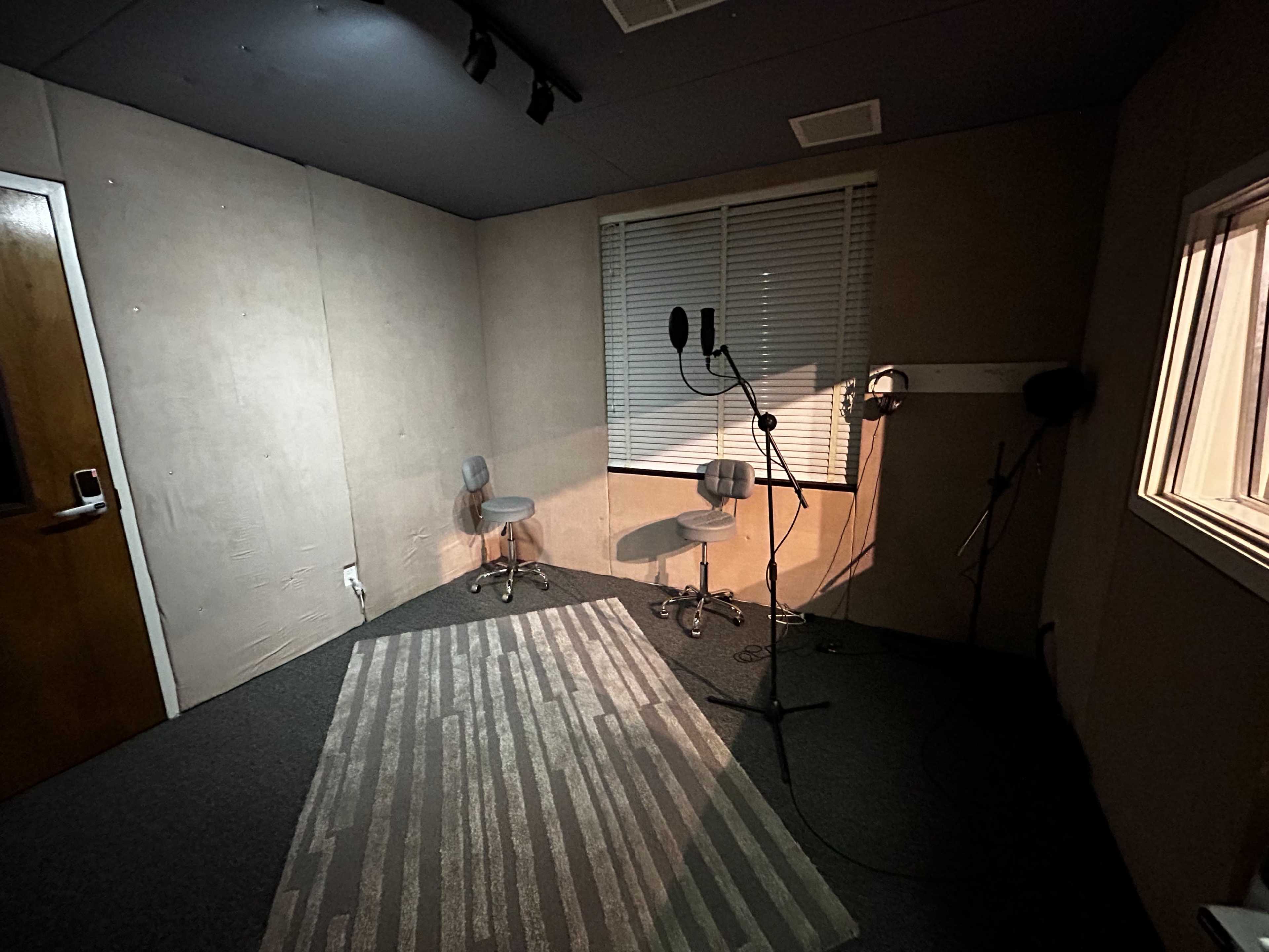 A small recording studio with two chairs, a microphone setup, and a window overlooking another room.