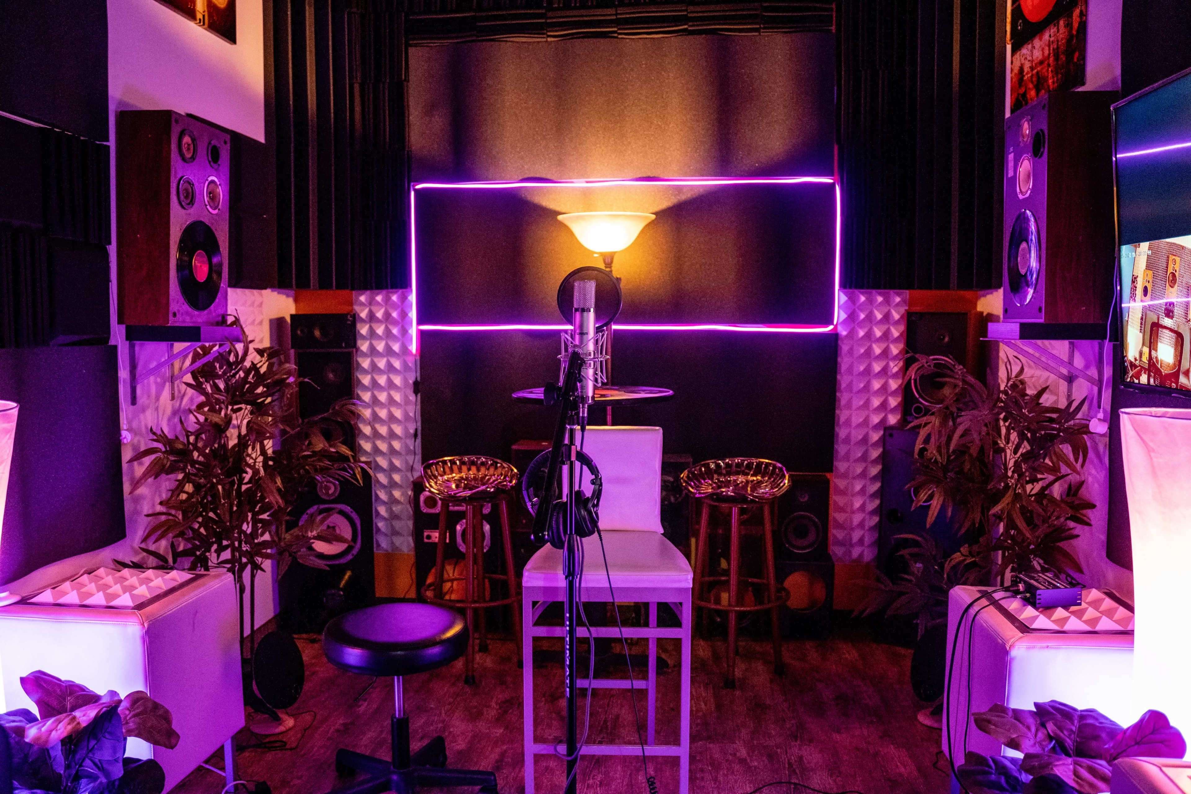 A sound studio featuring a microphone in front of a neon-lit wall, surrounded by bar stools and acoustic panels.