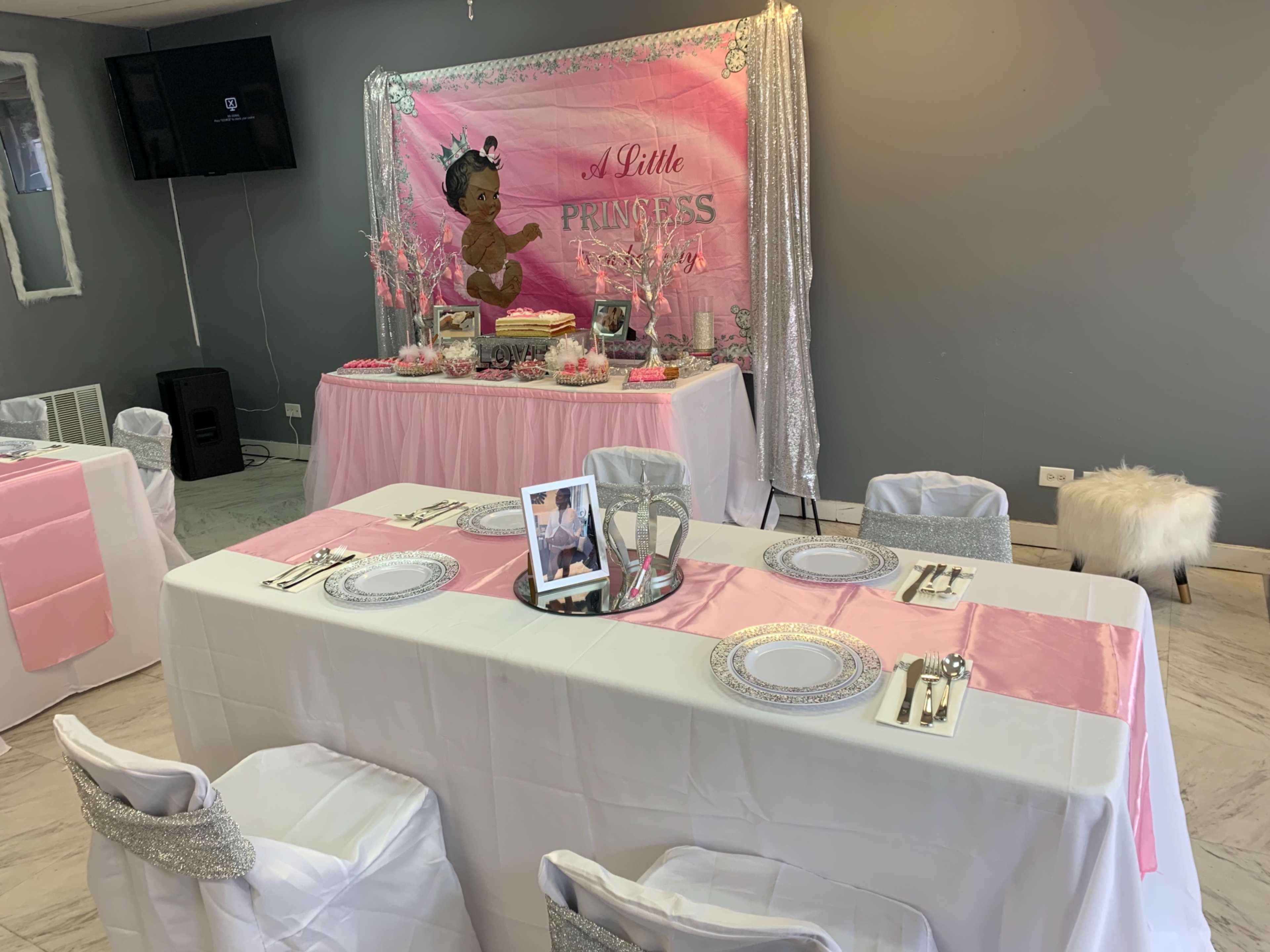 A decorated event space with two tables set for a party, featuring a pink theme and a display table adorned with a cake and decorations labeled "A Little Princess."