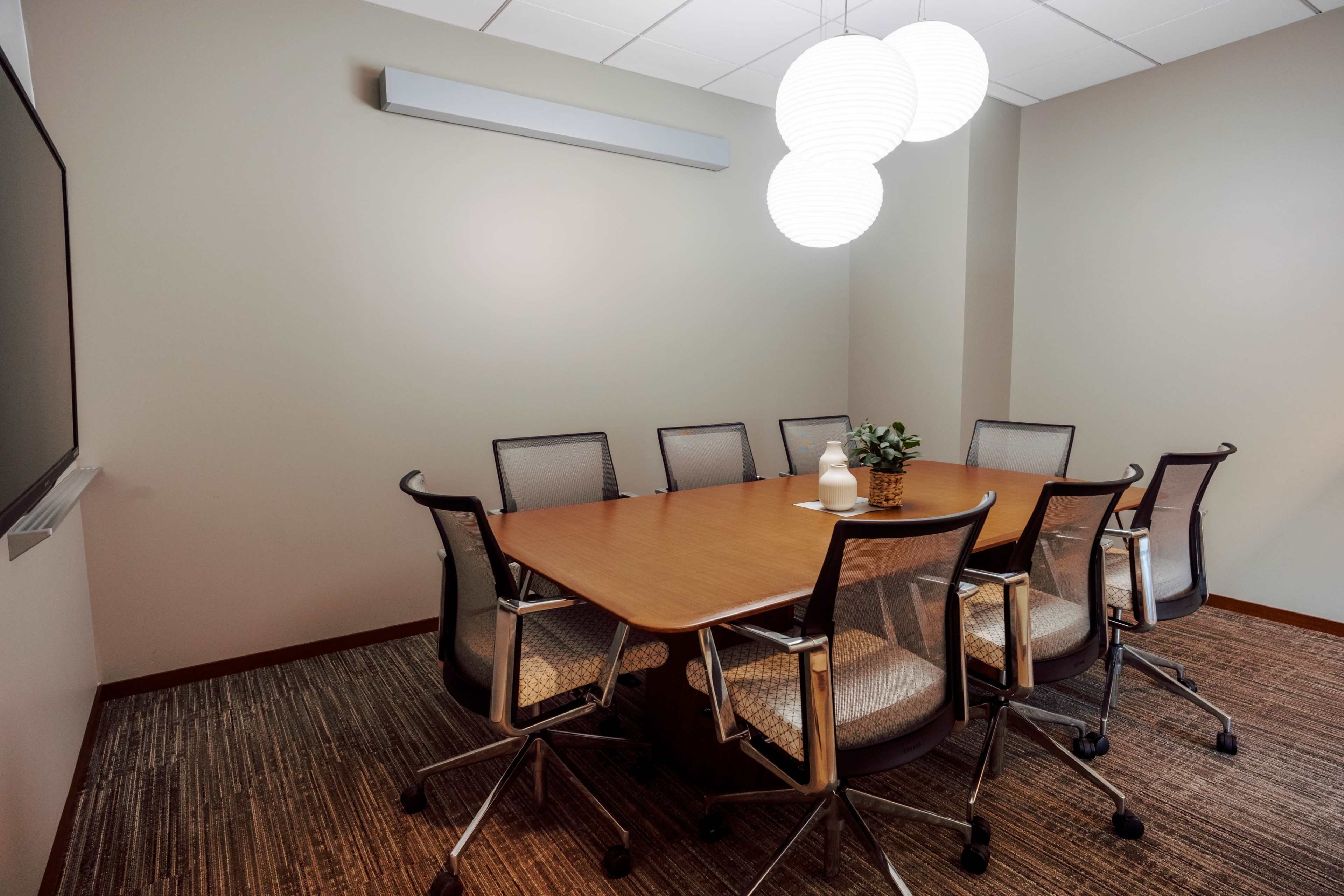A conference room features a large oval table surrounded by six mesh-backed chairs, illuminated by three pendant lights and equipped with a wall-mounted screen.