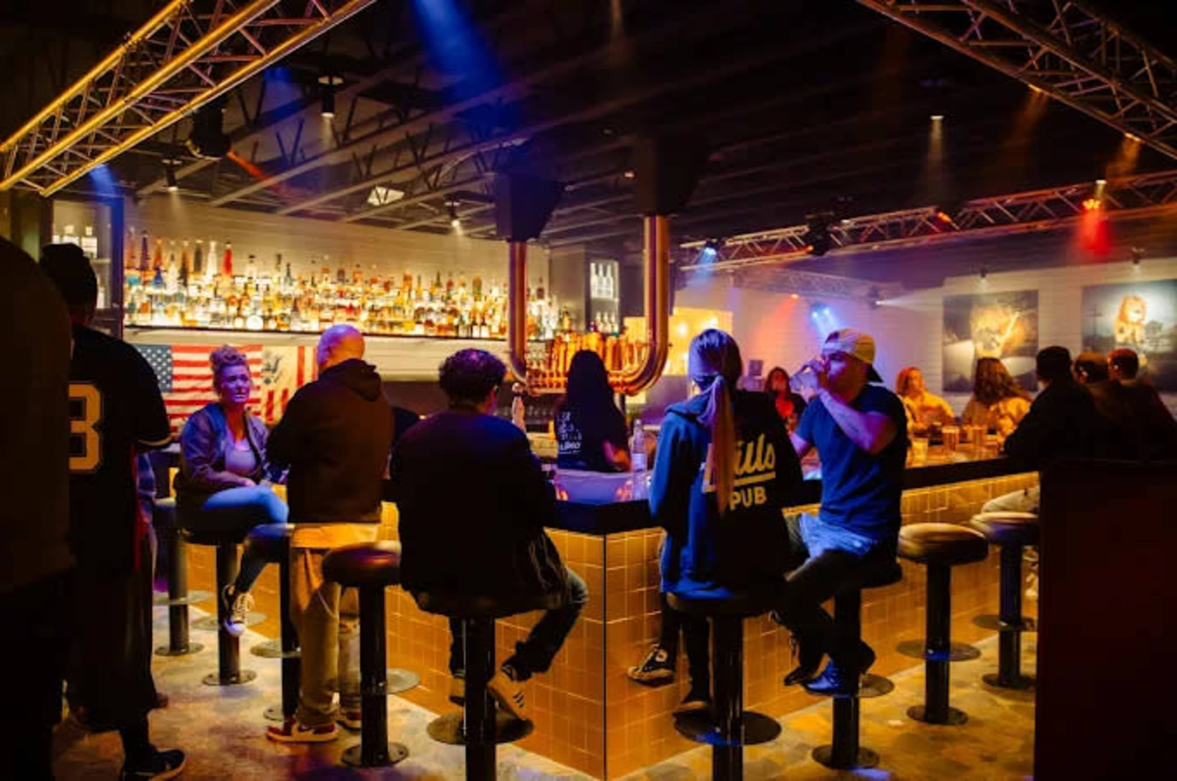 A dimly lit bar scene features patrons seated at a long counter with a variety of bottles displayed on shelves in the background.