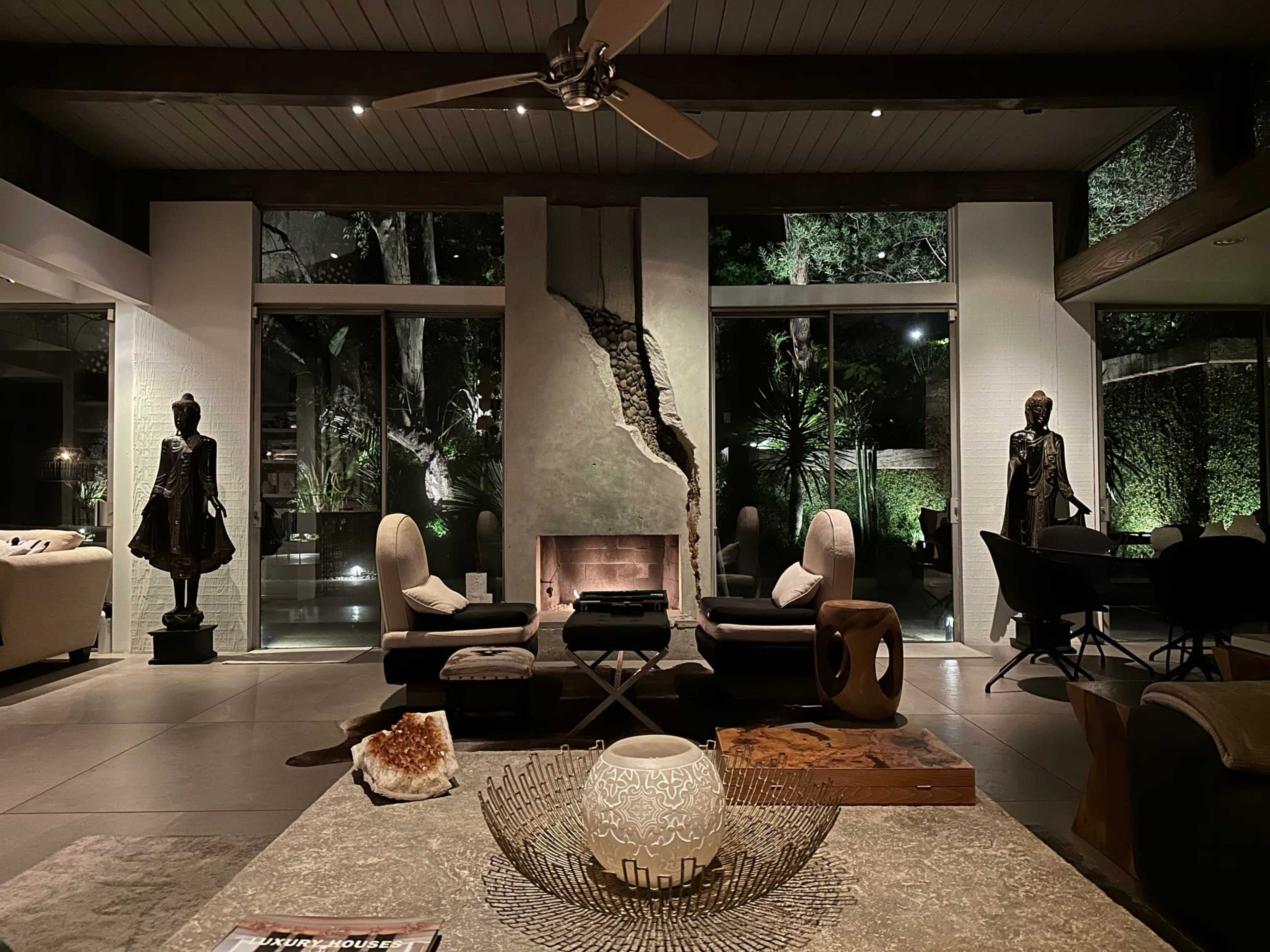 A modern living room with large windows overlooking a garden, featuring stylish furniture, decorative sculptures, and a central coffee table.