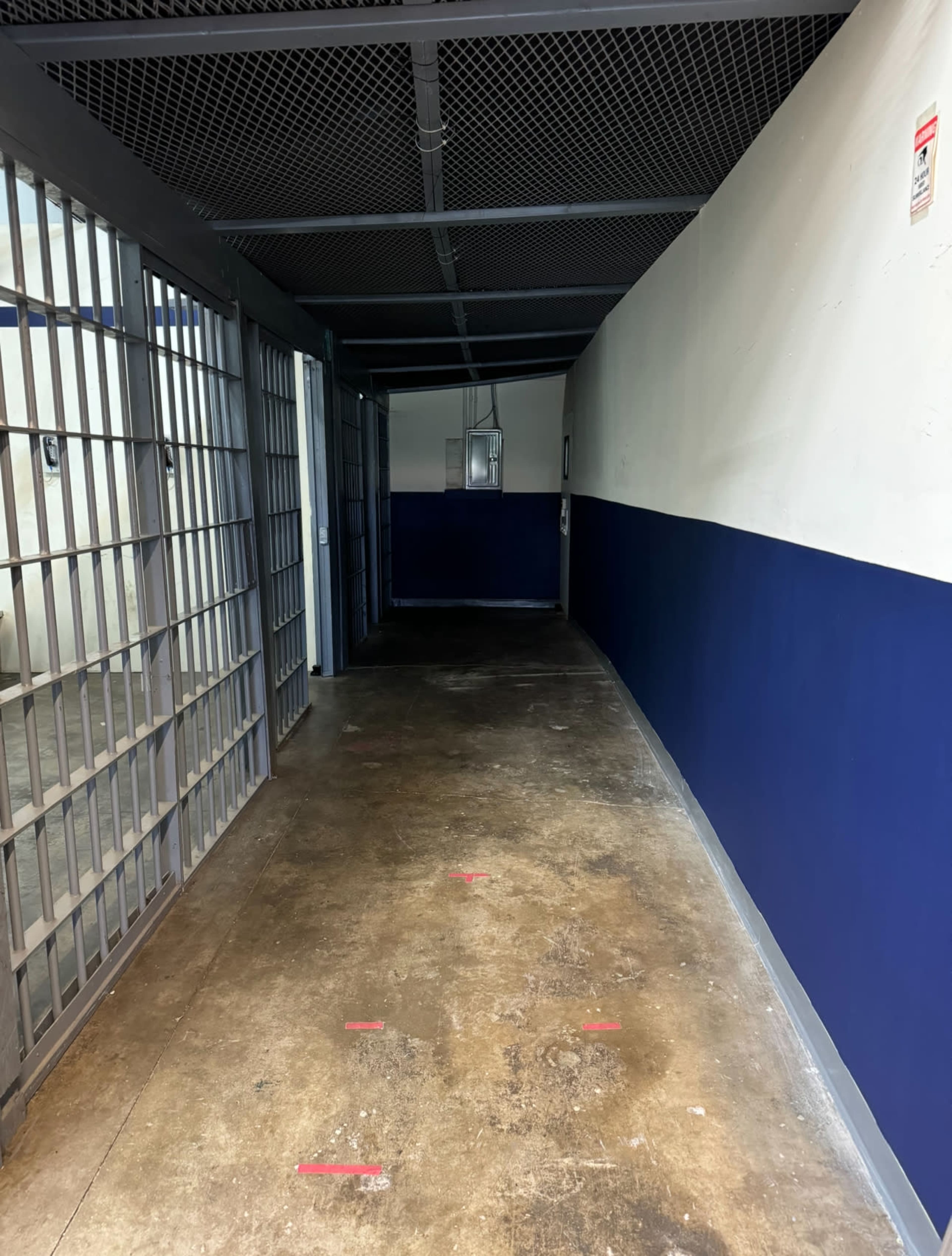 A narrow, dimly lit corridor lined with barred cells on one side and a blue-painted wall on the other.