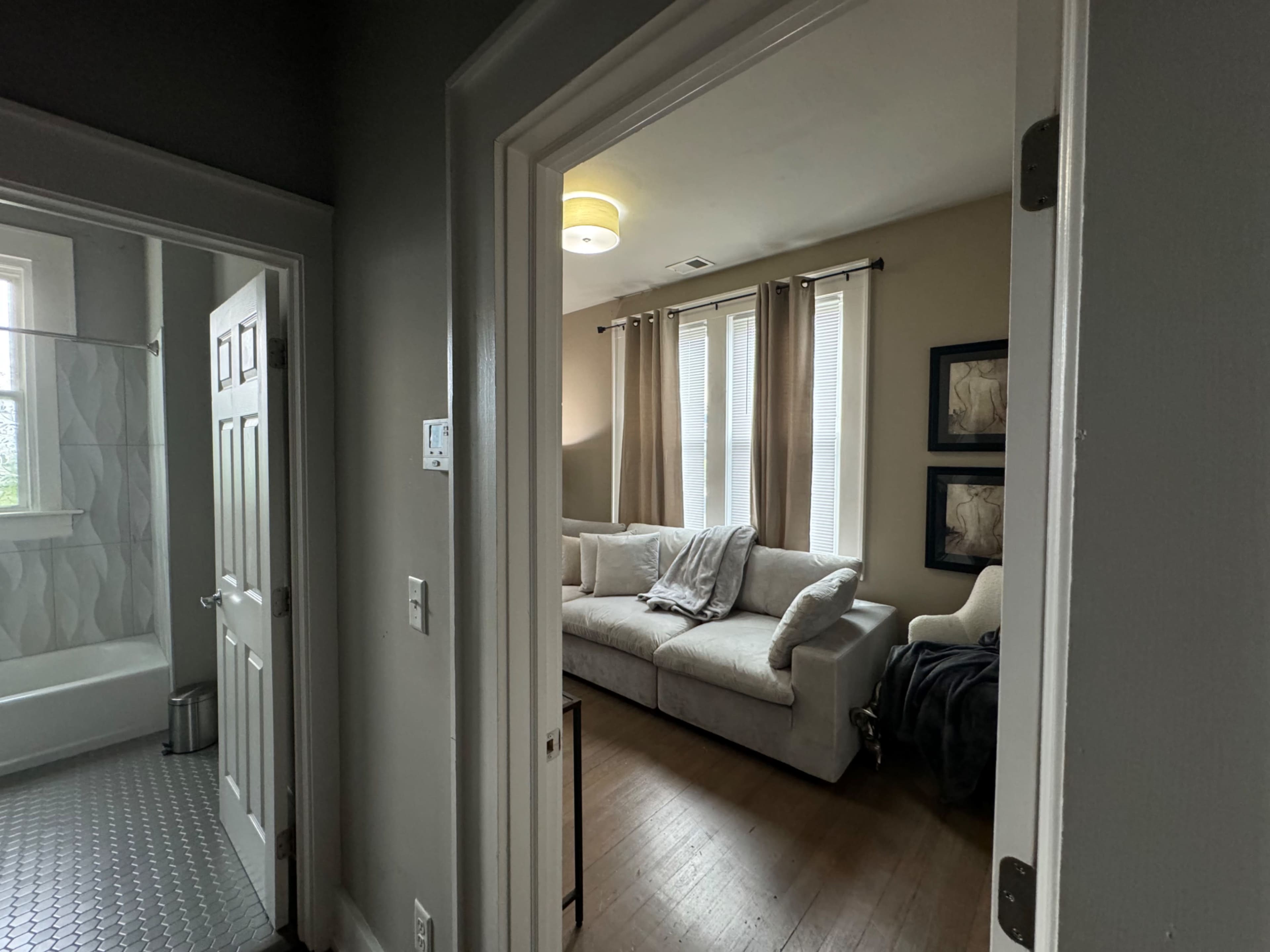 A view of a living room from a hallway, with a couch, armchair, and framed pictures on the wall, alongside a doorway leading to a bathroom.