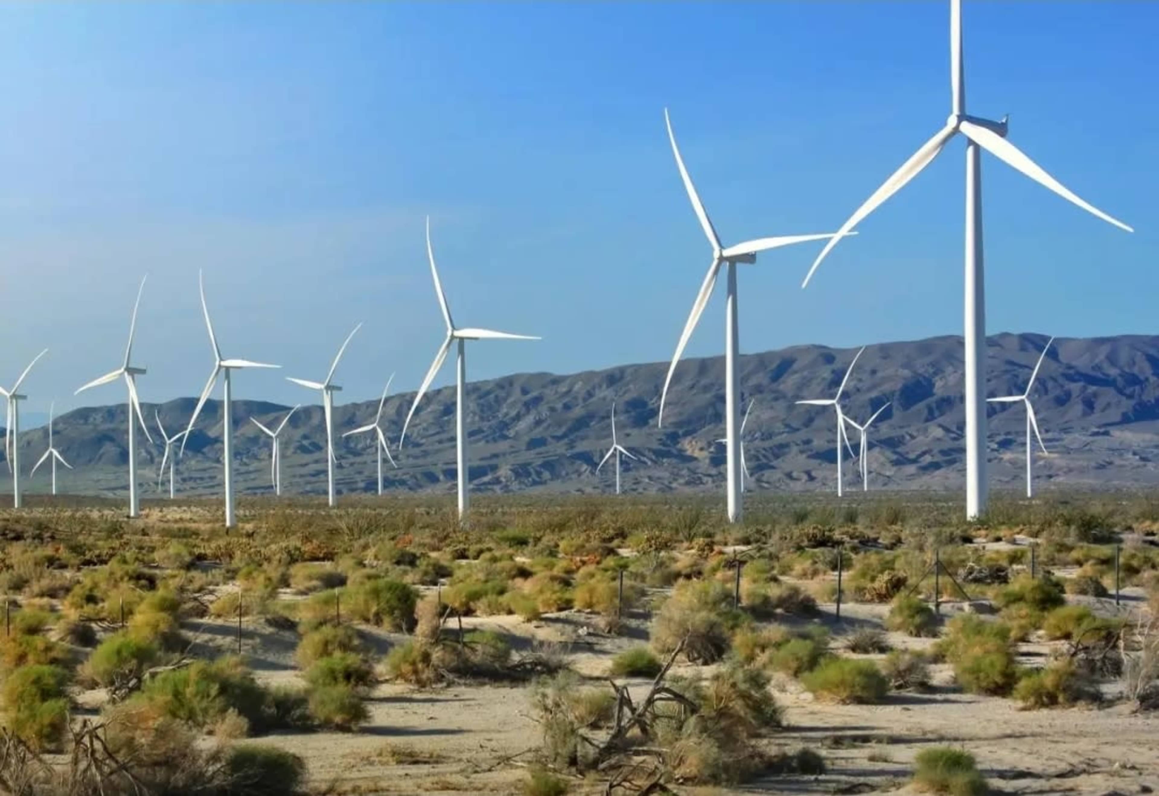 A row of large wind turbines stands on arid land with scattered vegetation and mountains in the background.