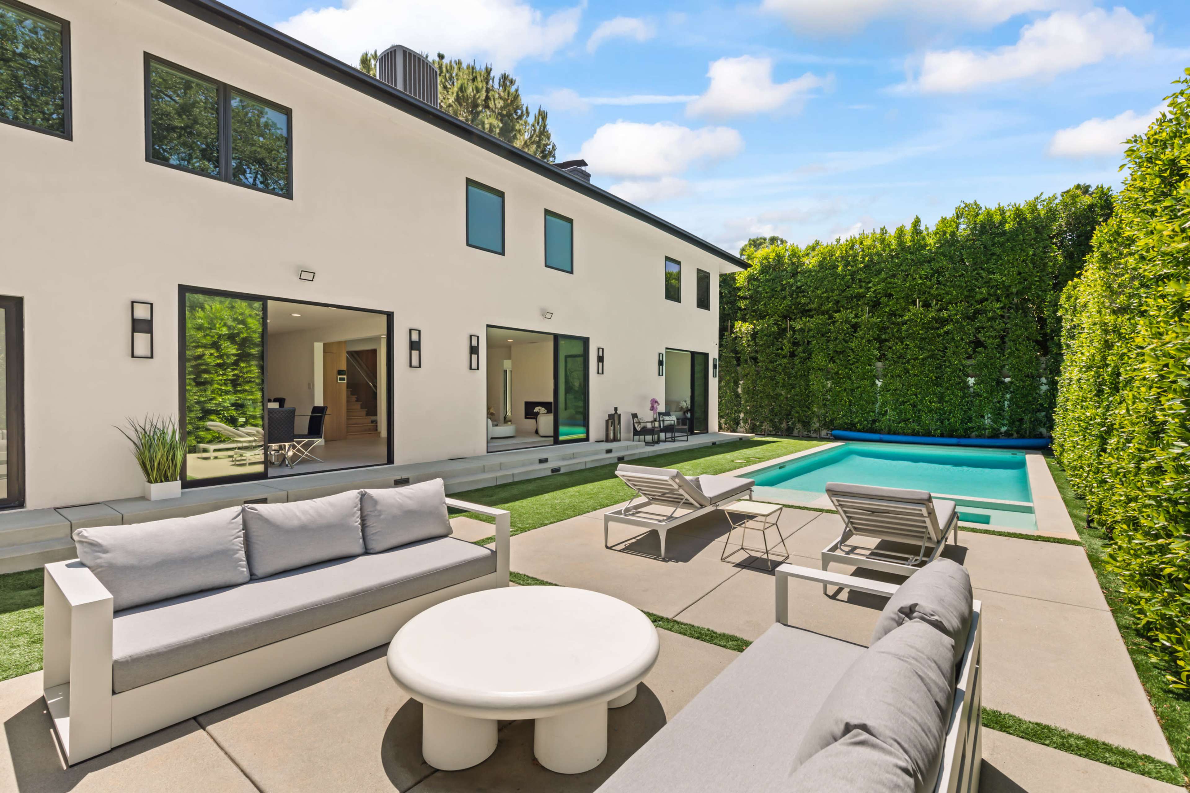 The image shows a modern backyard with a pool, surrounded by lush greenery, featuring outdoor seating and a spacious aesthetic.