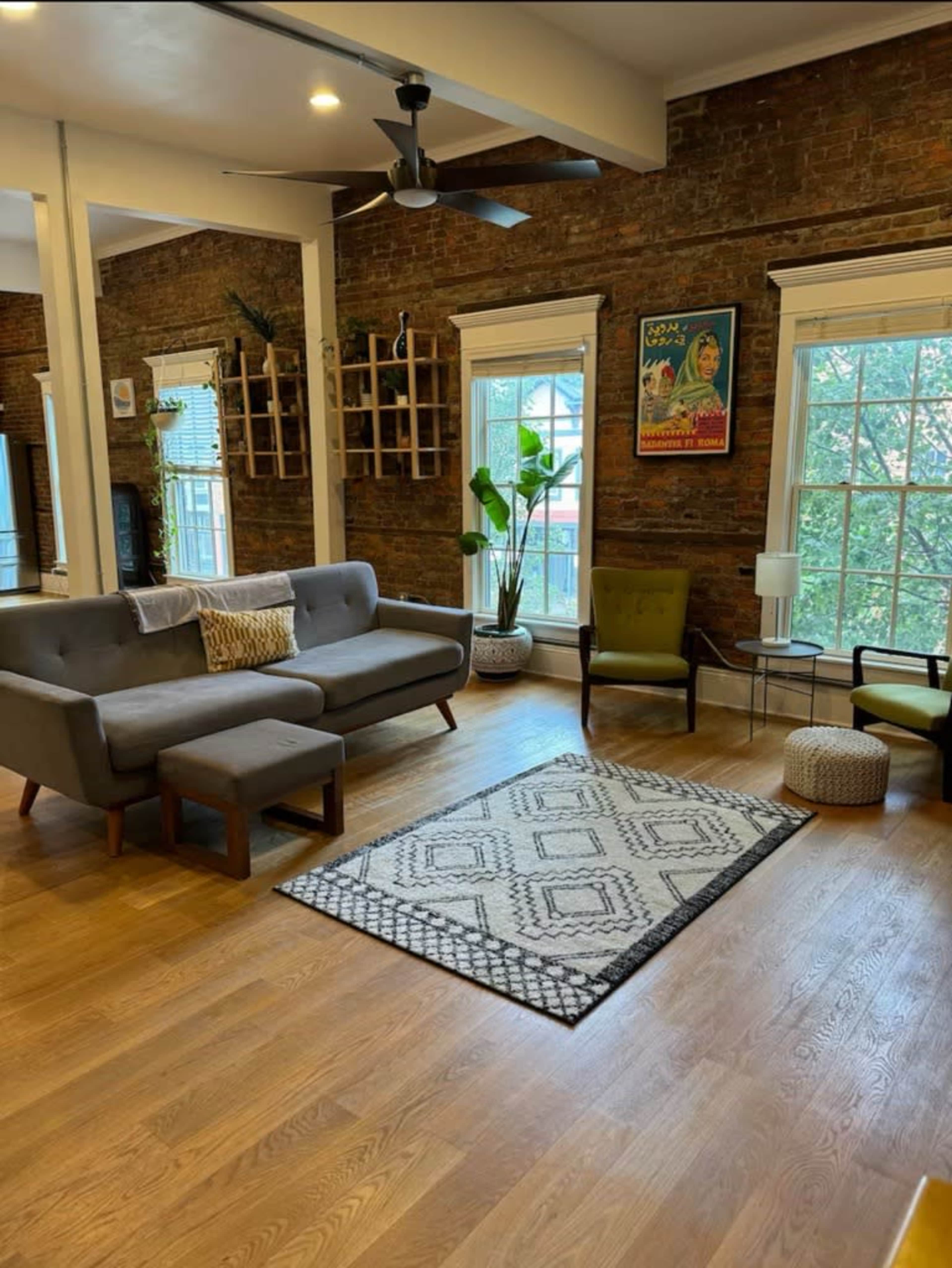 The image shows a spacious living room with exposed brick walls, a gray sofa, two green chairs, a patterned rug, and large windows providing natural light.