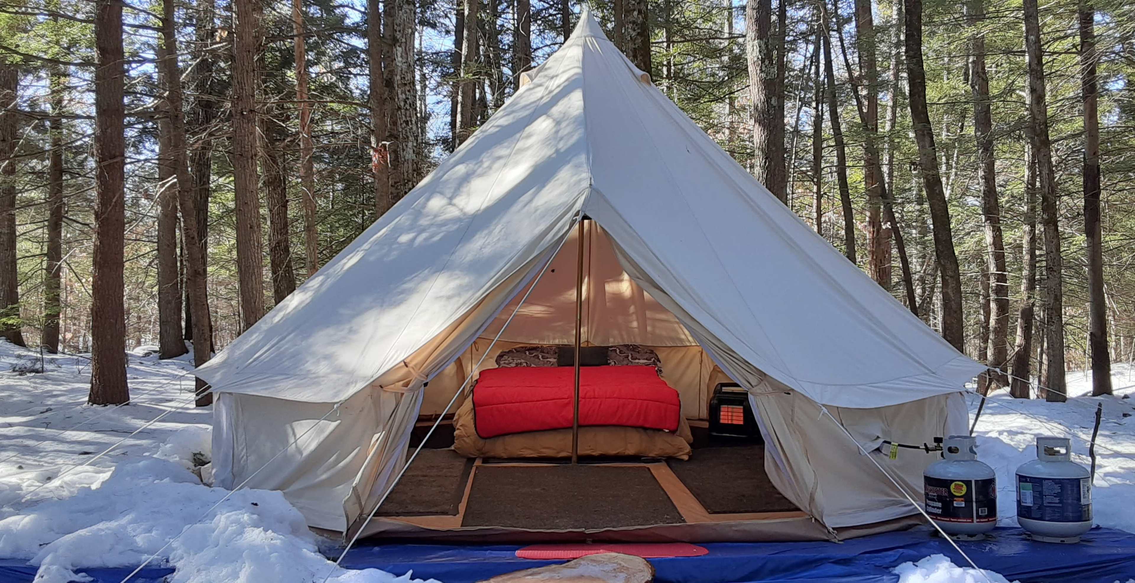 A large white tent is set up in a snowy forest, with a red bed inside and a propane tank outside.