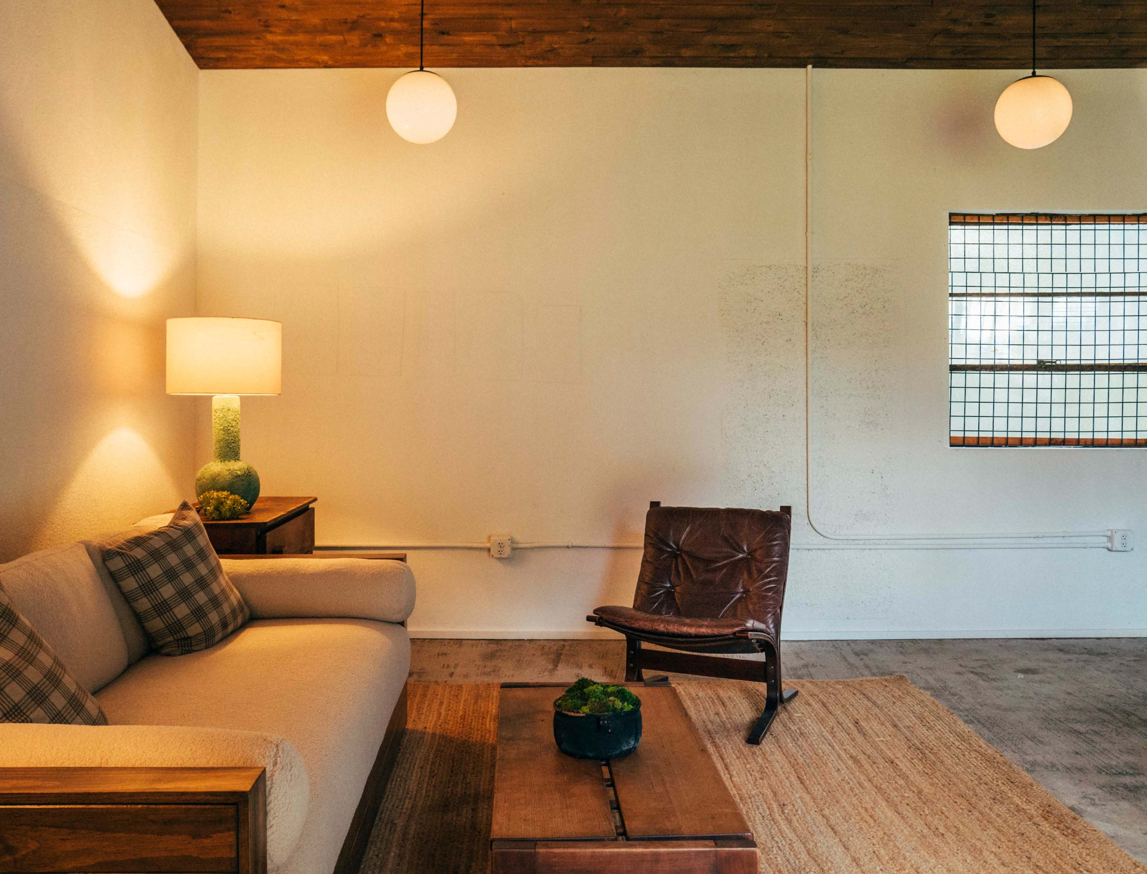 The image shows a minimalist living space with a light-colored sofa, a wooden coffee table, a potted plant, and a leather chair beside a lamp and a window.