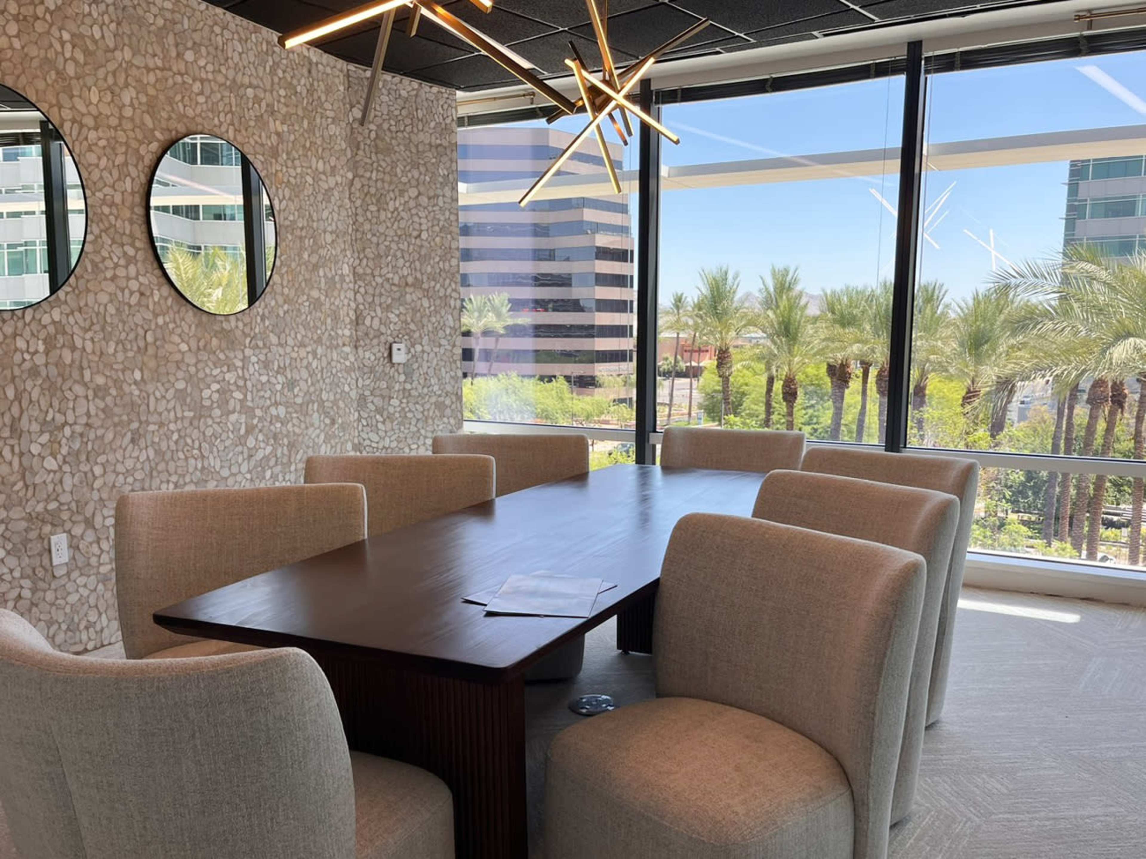 A modern conference room features a large wooden table surrounded by upholstered chairs, with large windows overlooking palm trees.