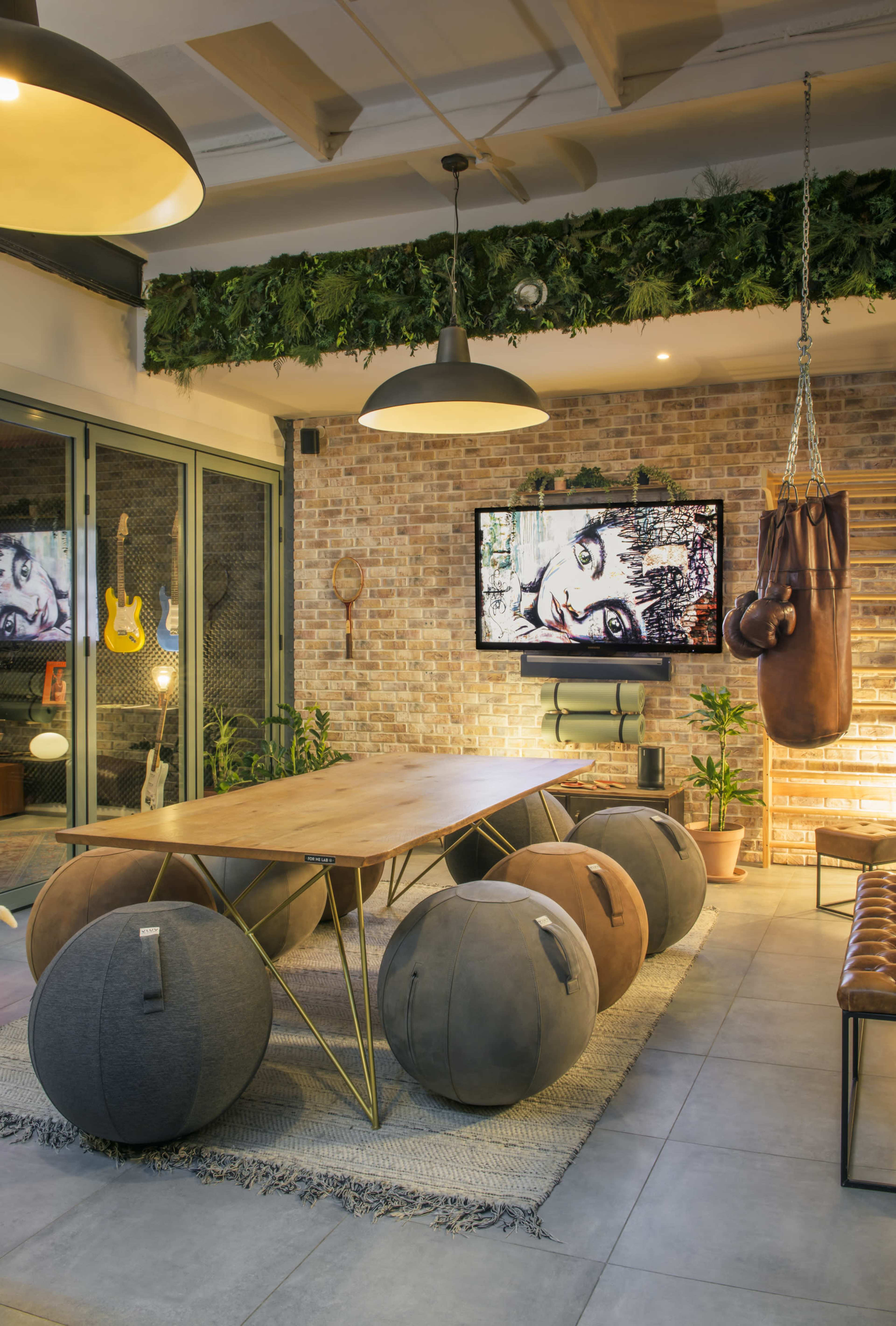 The image depicts a modern living space featuring a wooden table surrounded by round upholstered stools, with a wall-mounted television displaying artwork, and a punching bag hanging nearby, set against a backdrop of exposed brick and greenery.