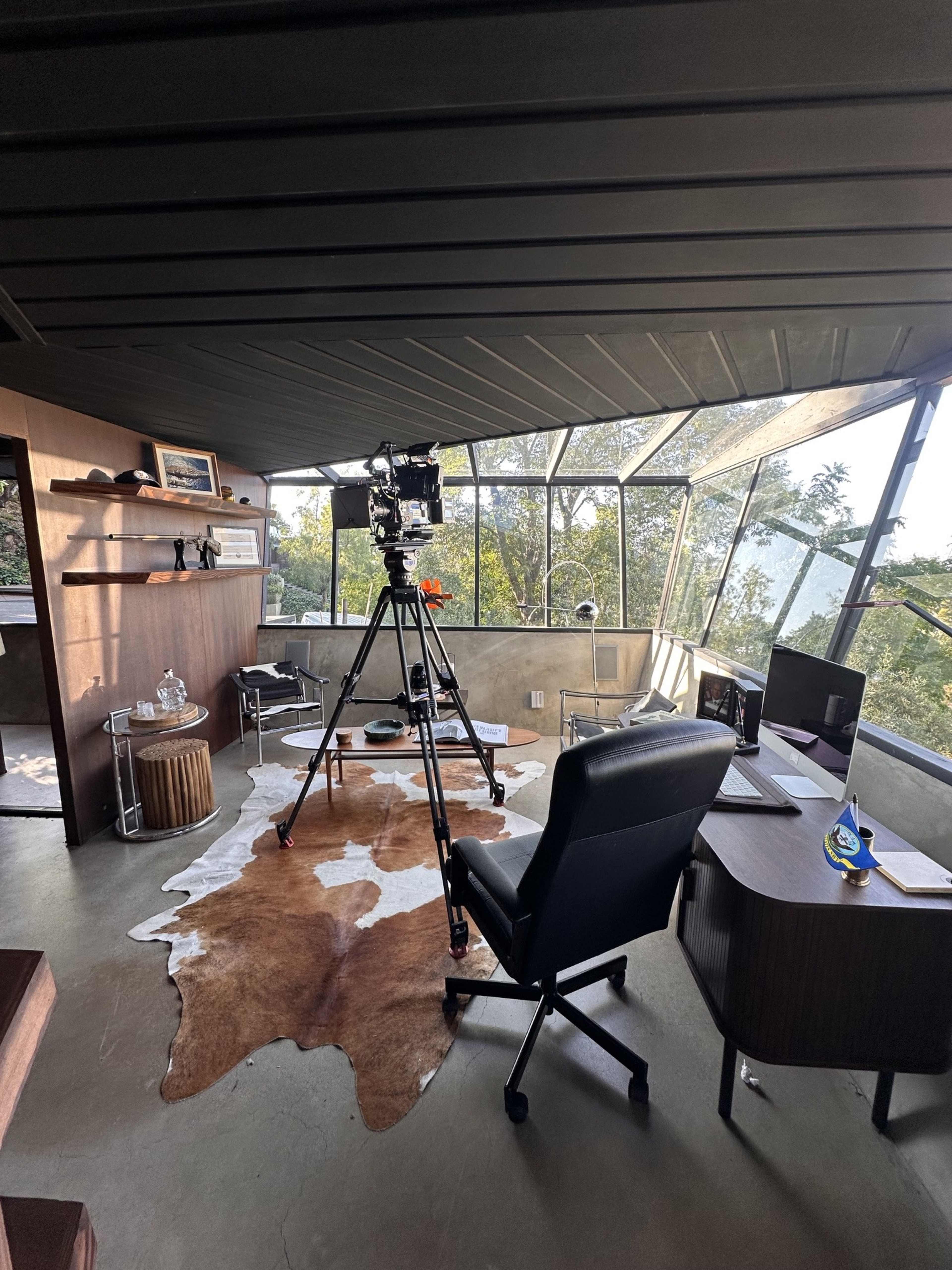 A modern office space features a camera on a tripod, a large window showcasing greenery, and a dark wood desk with a computer chair.
